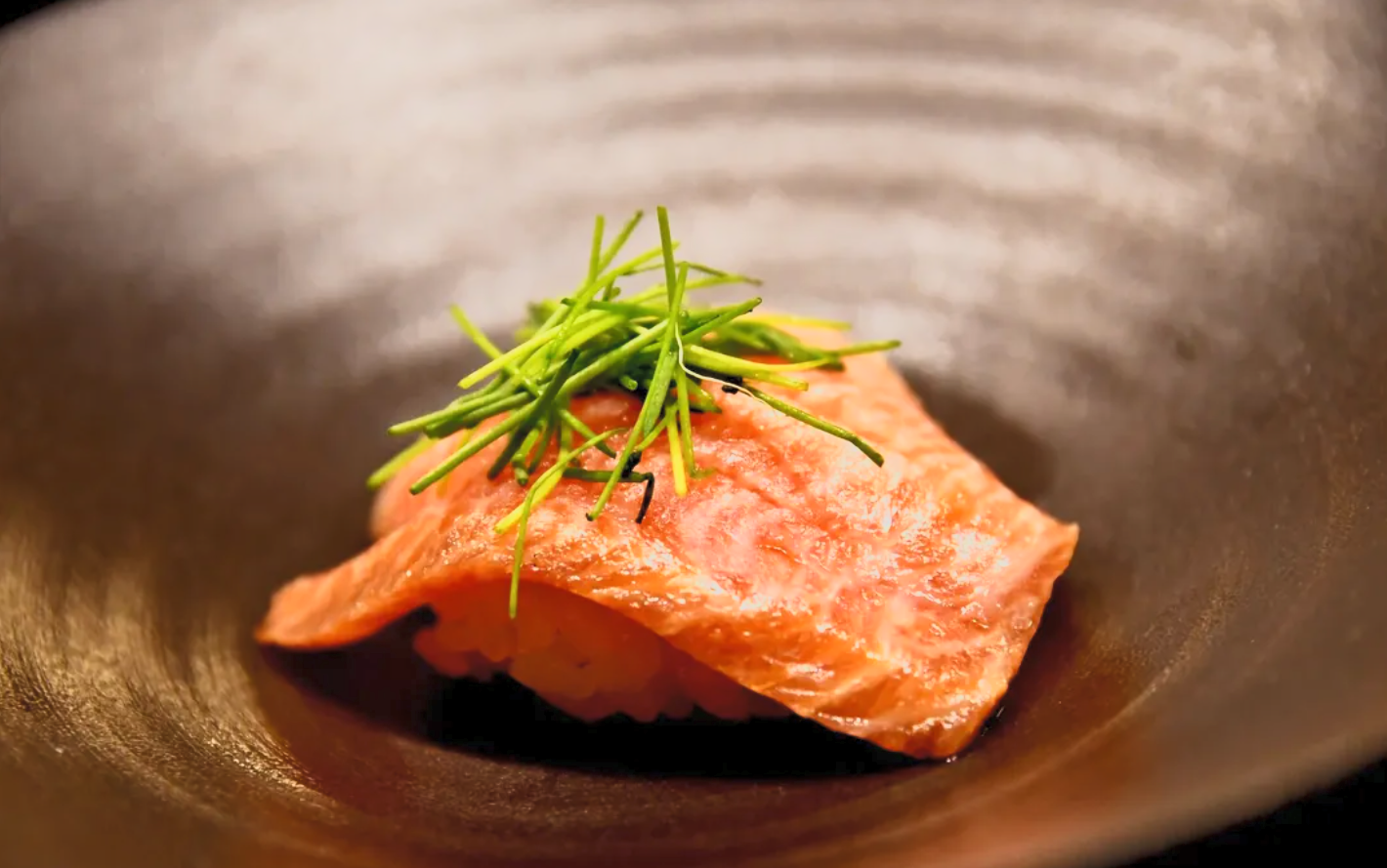 Close-up of a piece of cooked salmon topped with fresh green herbs, placed in a dark, textured bowl.
