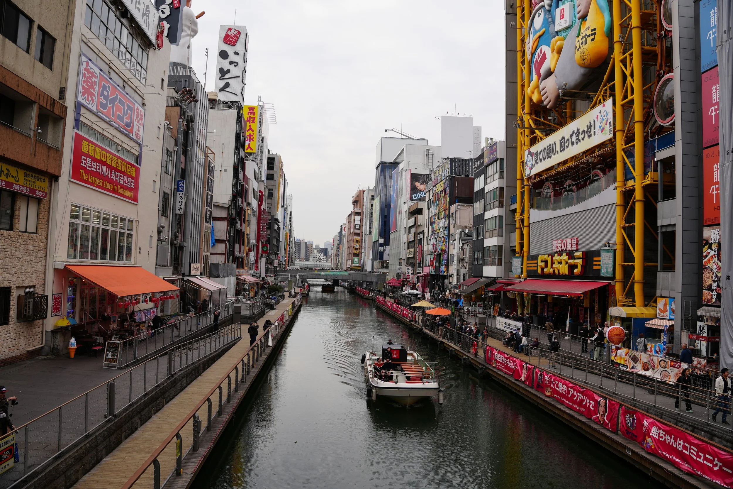 A bustling city canal scene with tall buildings, colorful billboards, and signs in Japanese. There are boats on the water, pedestrians on the walkway, and a large yellow structure with giant cartoonish feet and colorful objects on the right side.