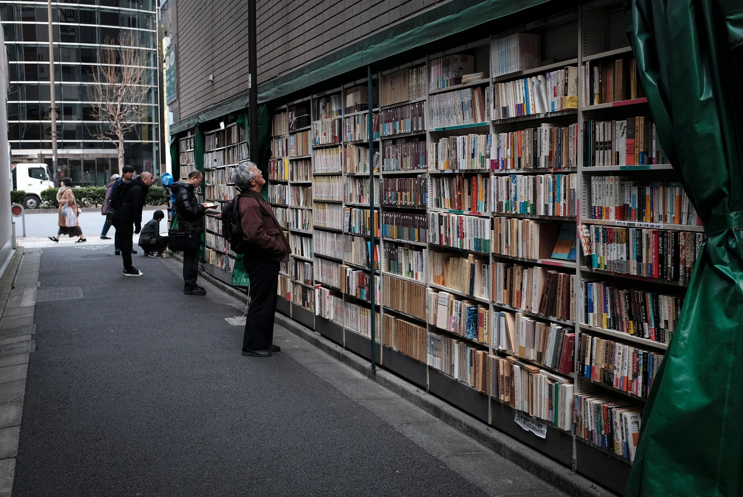 People browsing books on an outdoor book display in an urban area with city buildings and a leafless tree in the background.