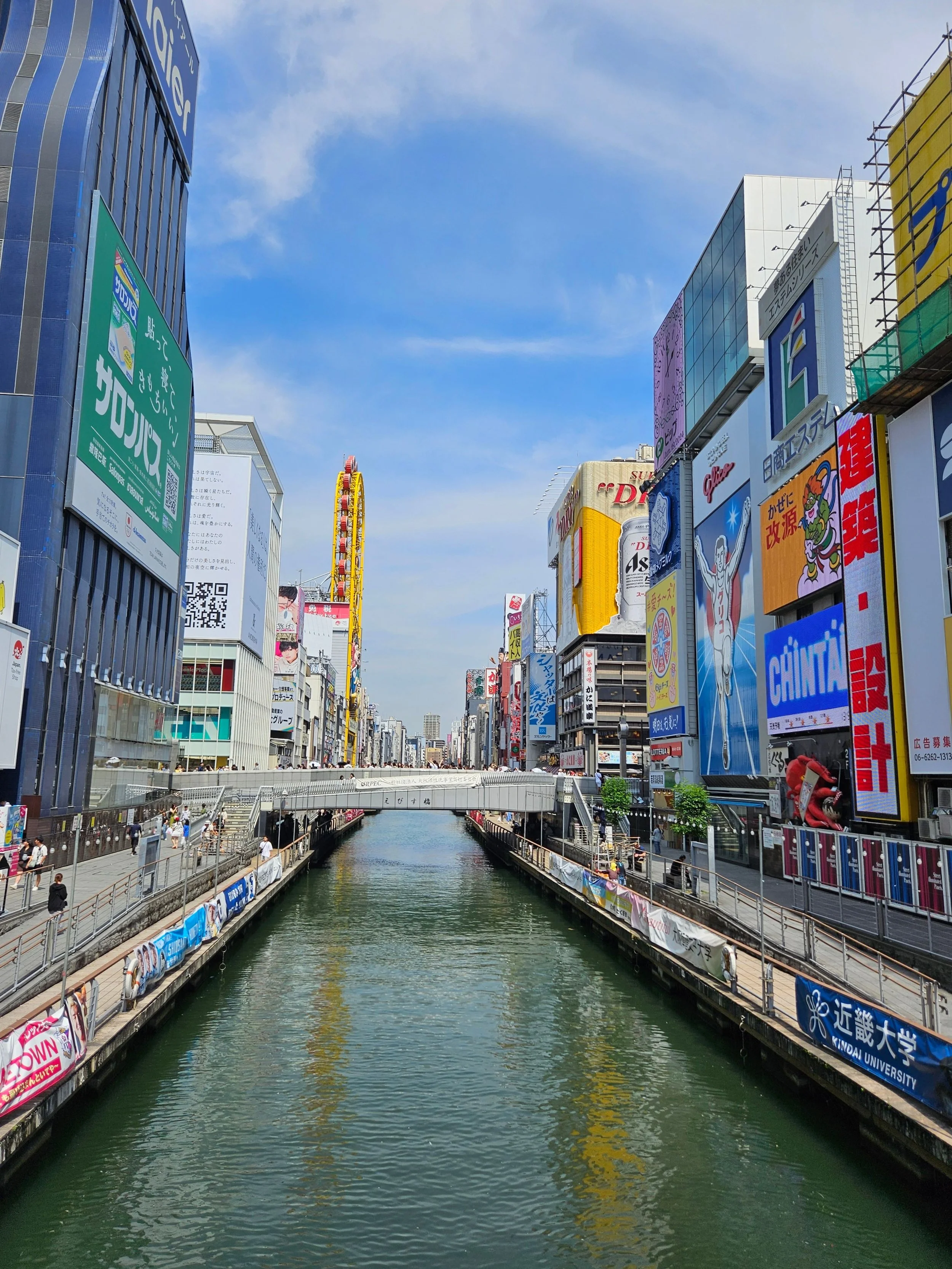 A cityscape with tall buildings on both sides of a canal, featuring numerous colorful billboards and advertisements, under a partly cloudy sky.