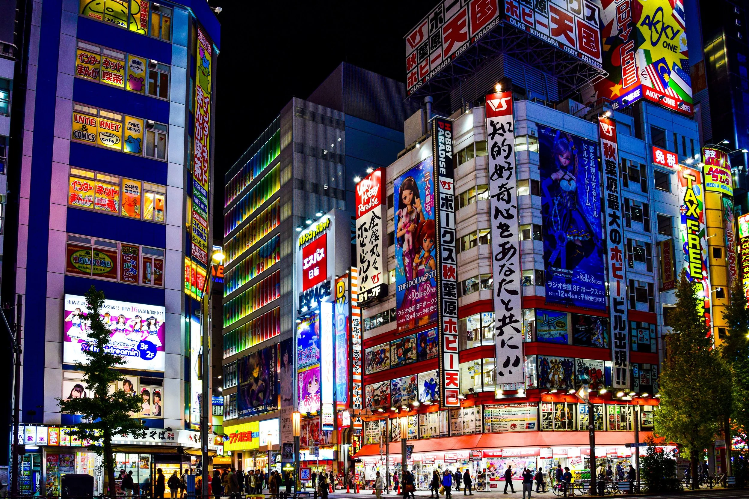 Night scene of a busy city with tall buildings covered in colorful illuminated advertisements and signs in Japanese, featuring anime characters, products, and brands, with people walking on the street below.