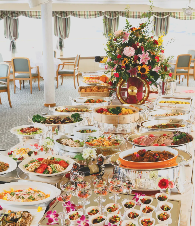 A buffet table with various dishes, a large floral centerpiece, and glasses of dessert, in a bright room with large windows and curtains.