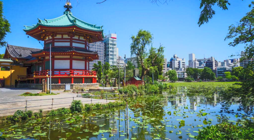 Traditional Japanese pagoda near a pond with water lilies and water plants, in an urban park setting, with modern buildings in the background under a clear blue sky.