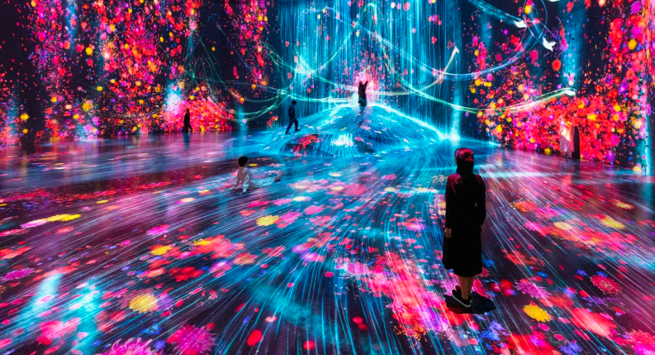 People experiencing an immersive digital art installation with colorful lights, projections of flowers, and dynamic patterns on the walls and floor.
