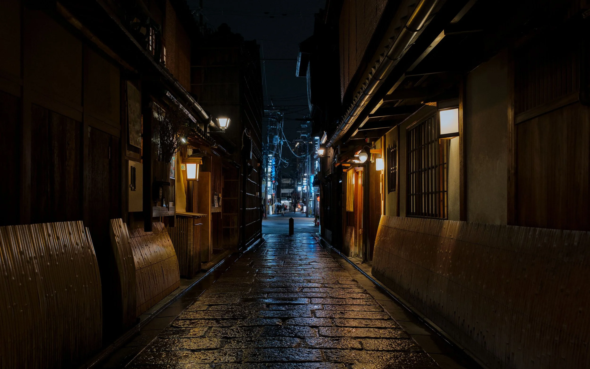 Night scene of a narrow alleyway with wet pavement, illuminated by wall-mounted lamps on surrounding buildings, with a view of city lights and power lines in the distance.