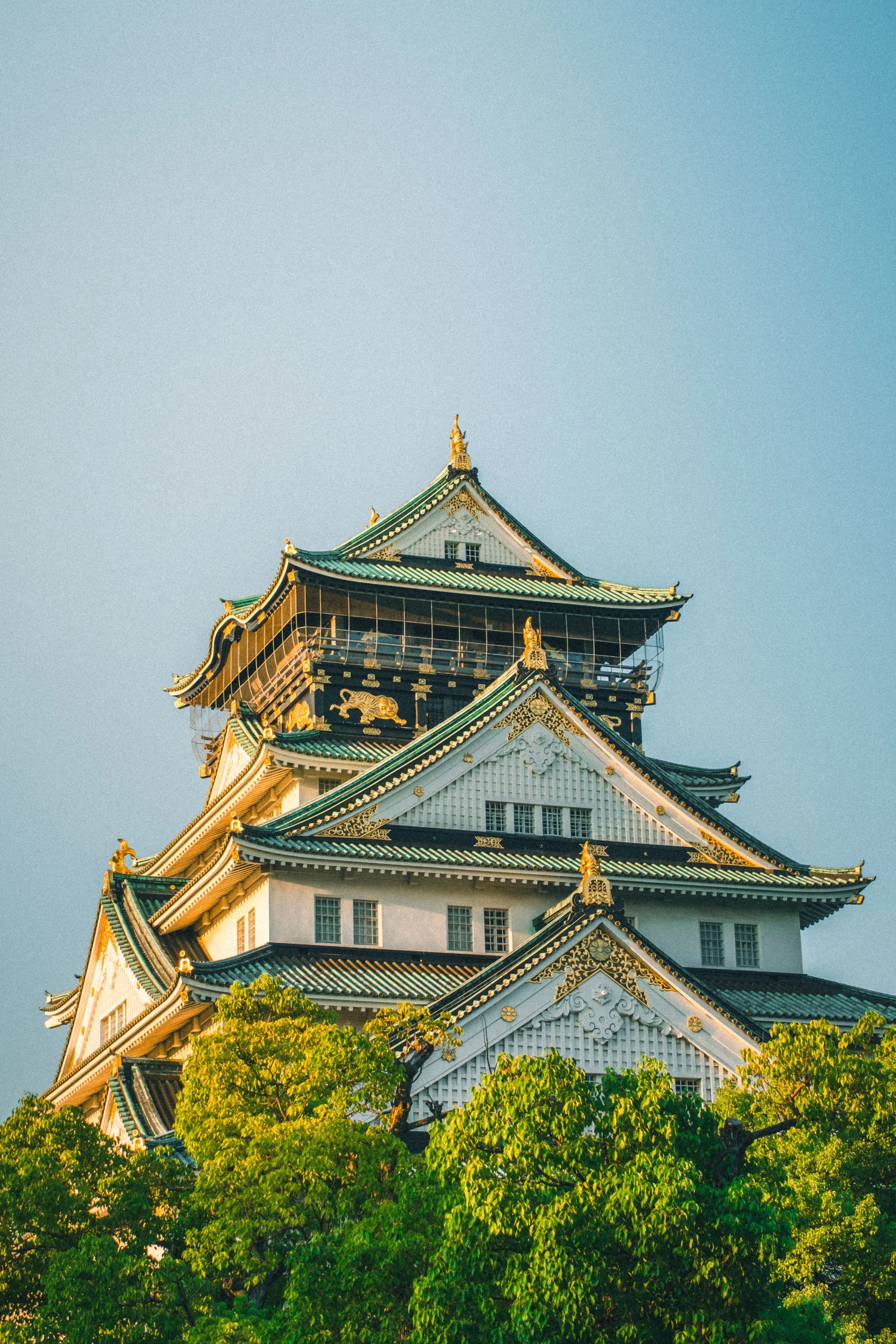 Japanese castle with green-tiled roofs, ornate gold accents, and surrounded by lush green trees, against a clear blue sky.