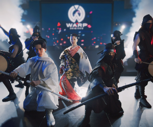 A group of performers on stage, dressed in martial arts attire, holding swords, with a background screen displaying the WARP logo and stage lights.