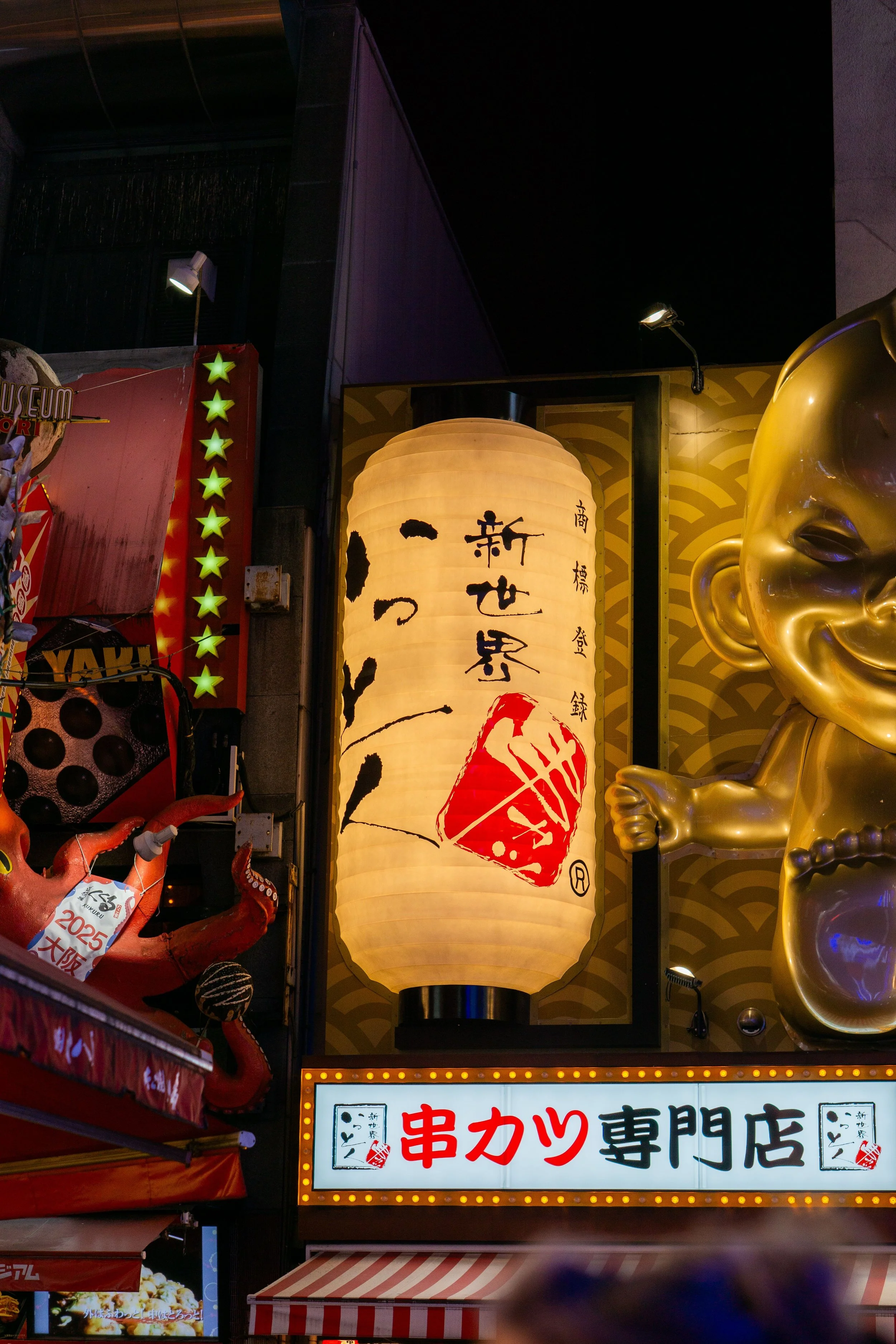 Street scene at night with illuminated signs, a large Japanese paper lantern, and a prominent golden Buddha head sculpture.