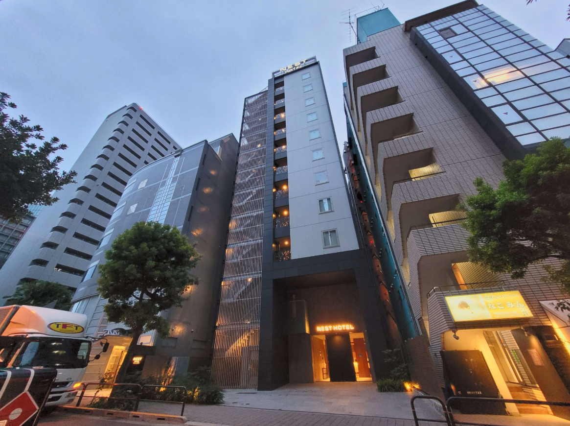 Tall modern hotel building with illuminated entrance and sign reading 'Nest Hotel' in an urban area, surrounded by other skyscrapers and trees.