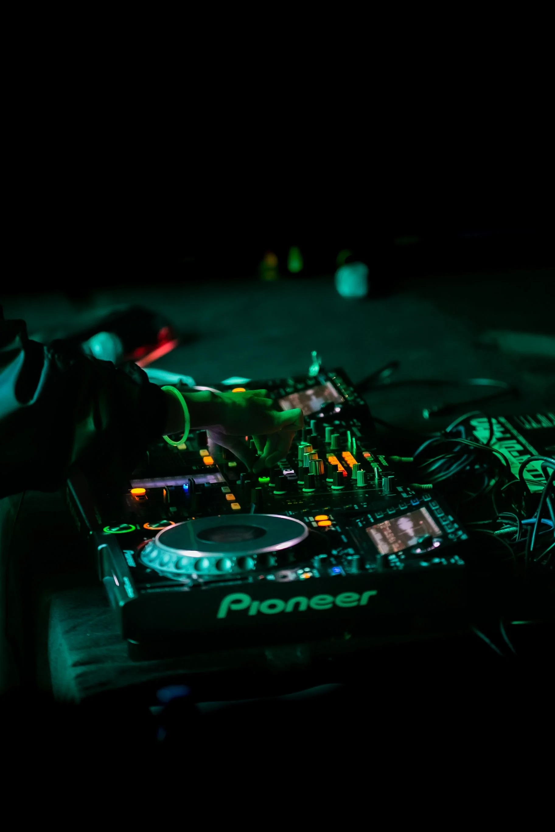 Nighttime scene of a DJ's hands operating a Pioneer DJ setup with colorful lights, surrounded by a dark environment and some blurry background objects.