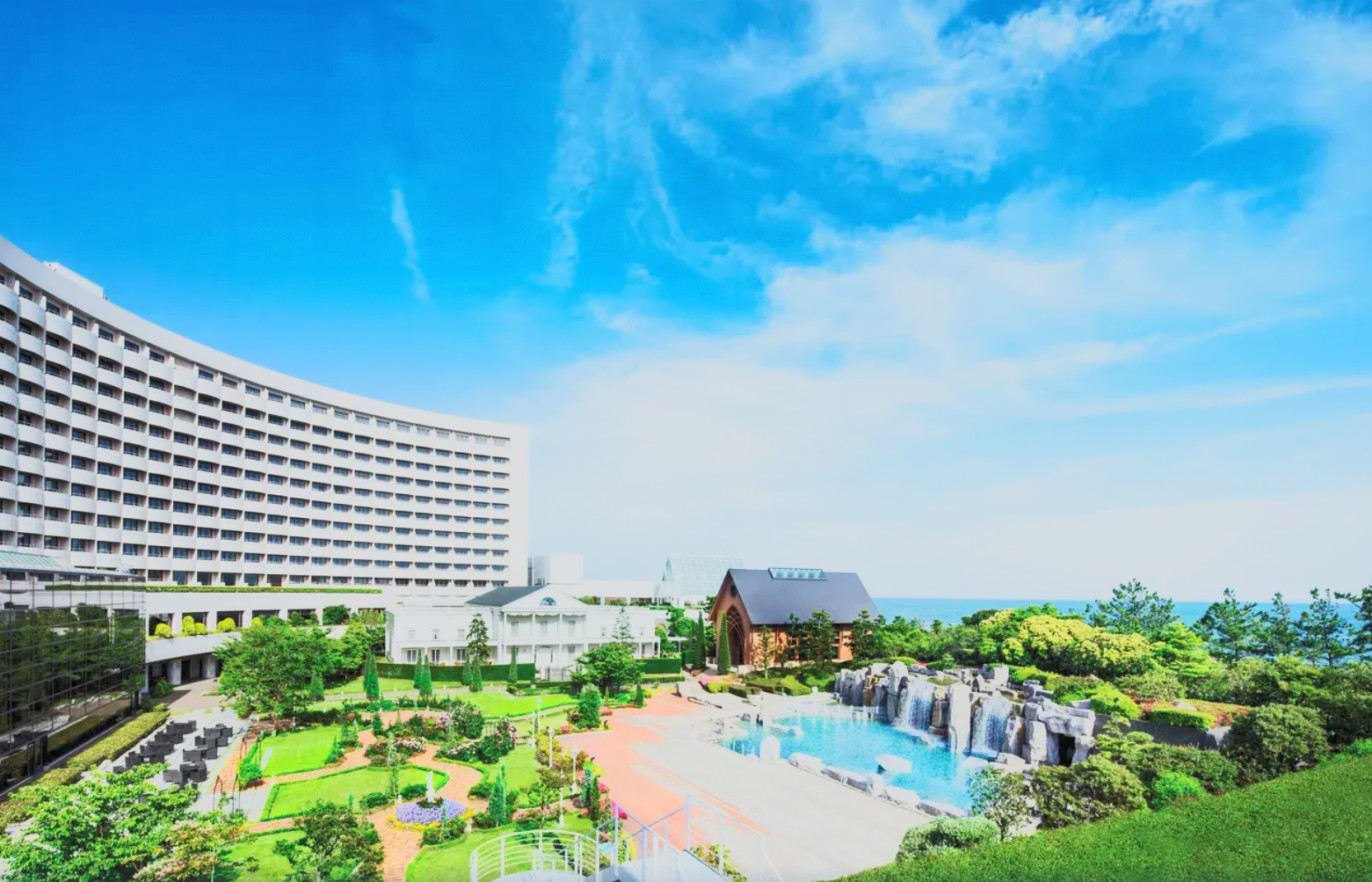 A large hotel building with a curved facade, a landscaped garden with trees and pathways, a fountain with waterfalls, and a clear blue sky with some clouds.