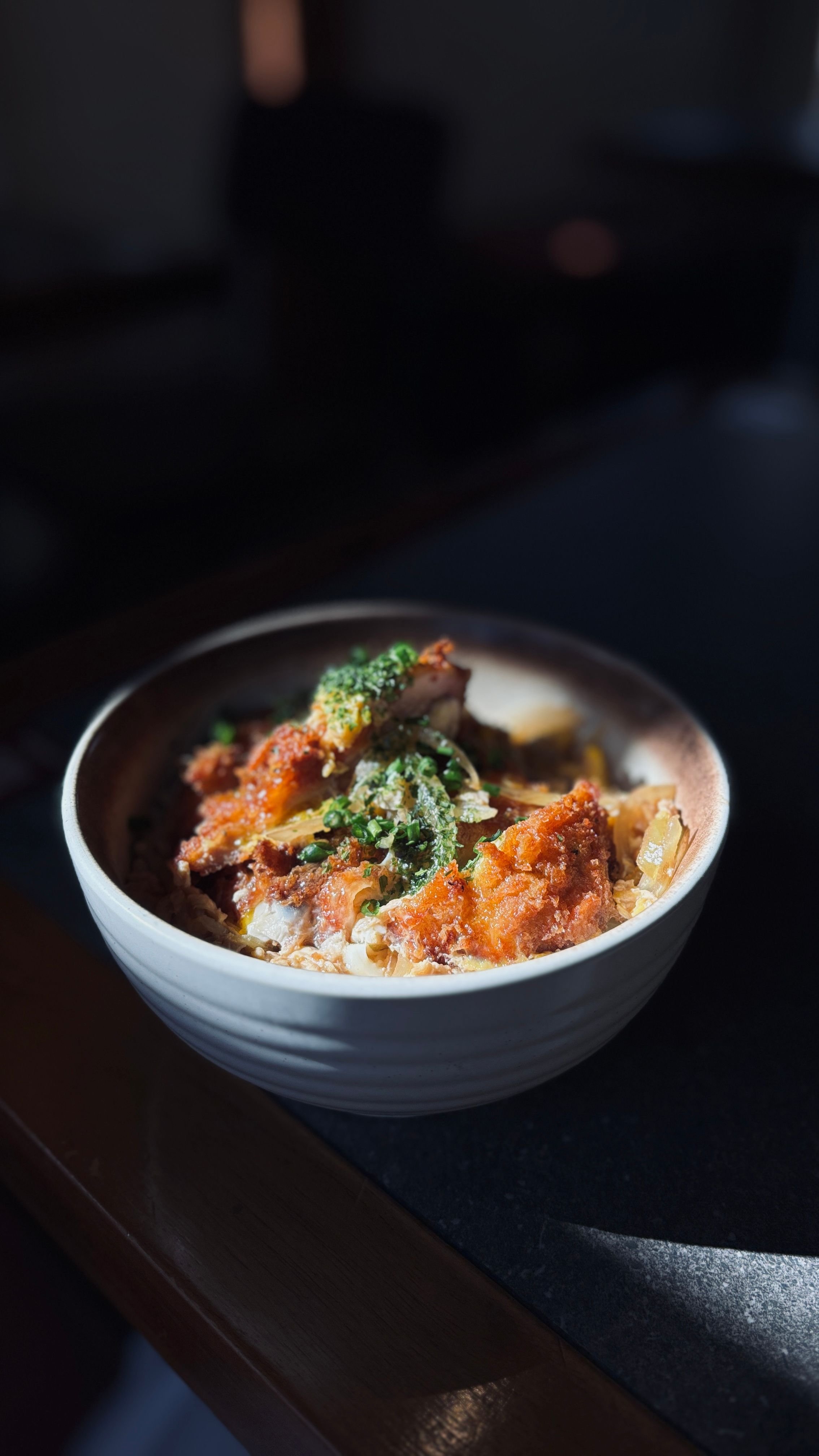 A bowl of baked dish topped with green herbs, possibly chopped scallions or chives, and other ingredients, sitting on a dark surface with a blurred background.
