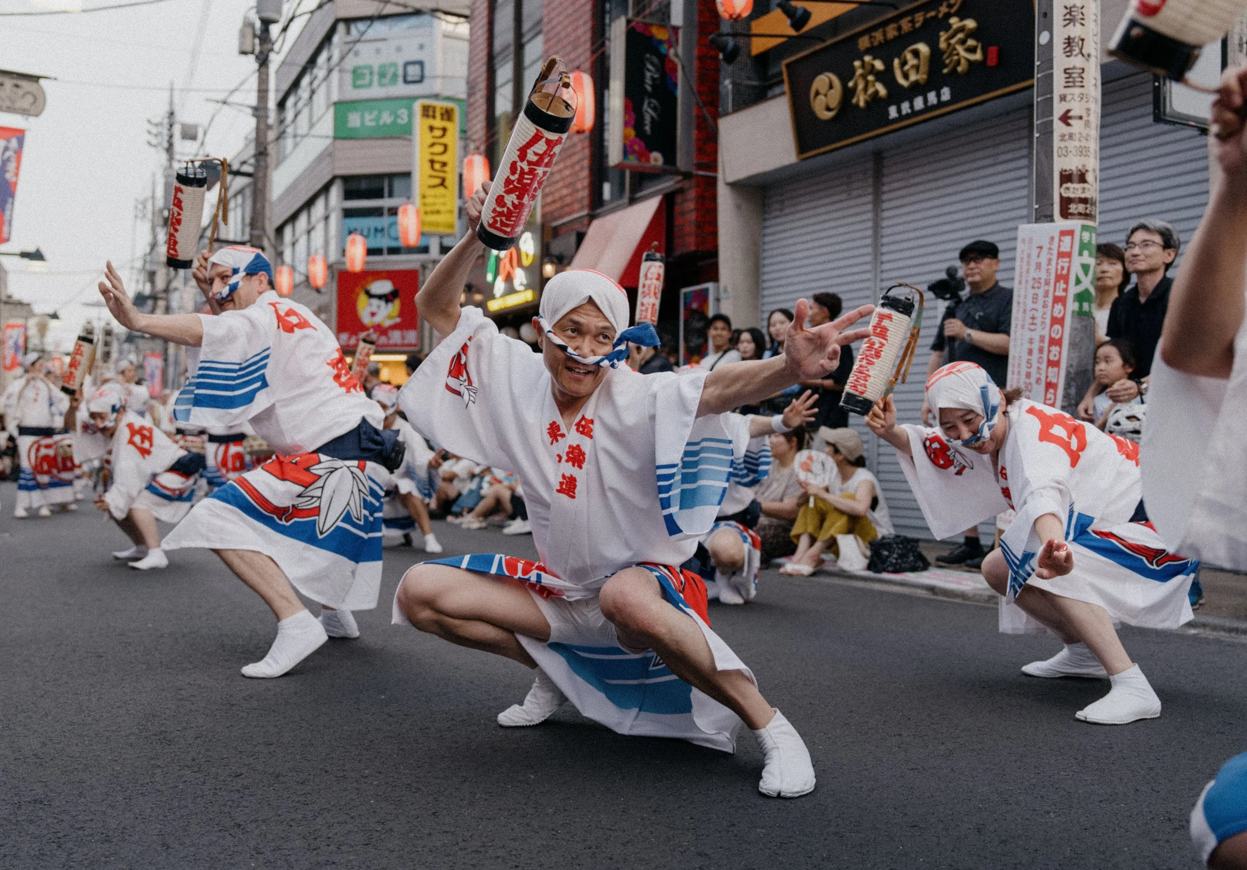 A group of people dressed in traditional Japanese festival attire participating in a cultural dance parade on a street, holding paper lanterns.