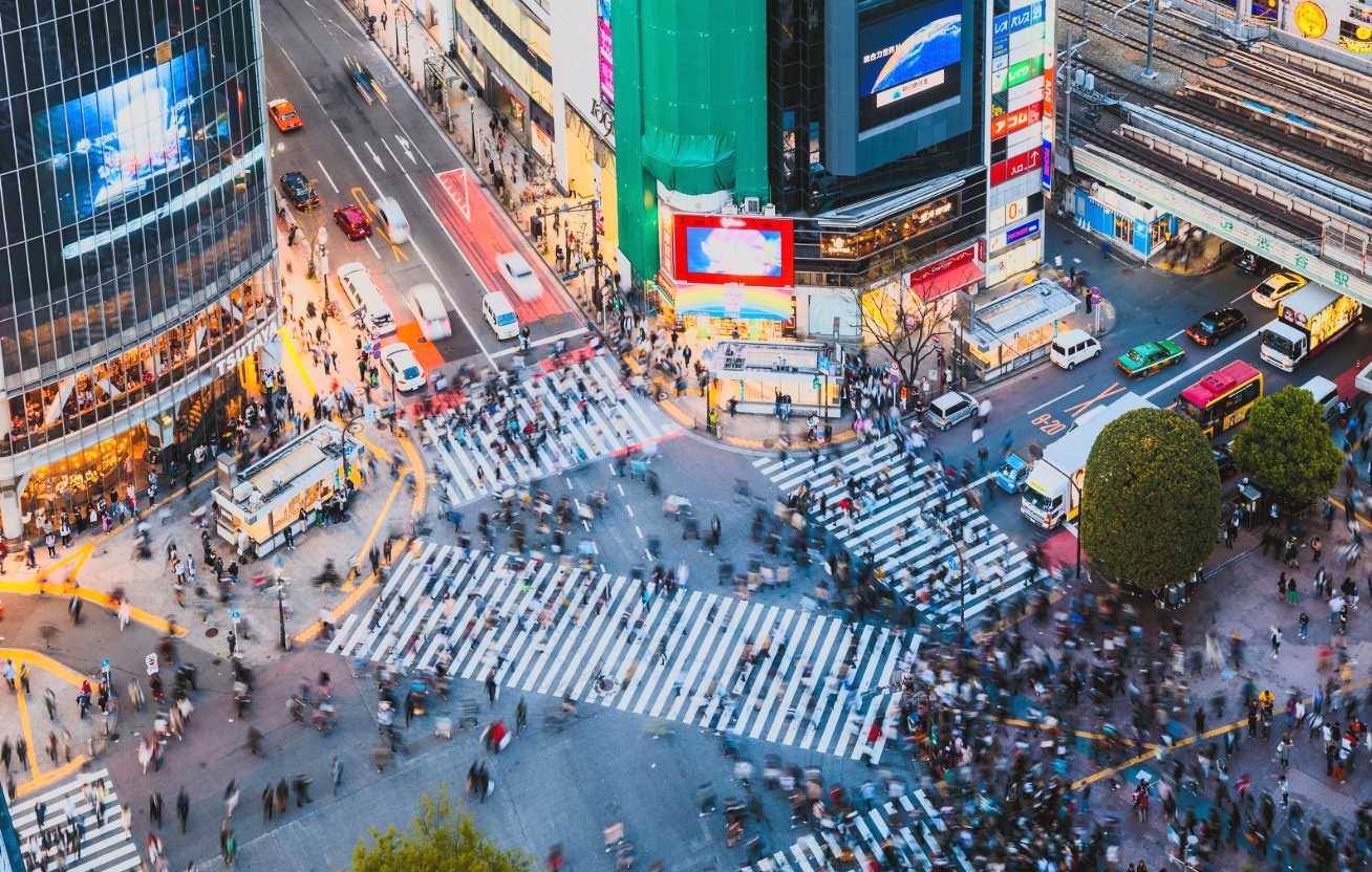Bird's eye view of a busy urban intersection in Tokyo, Japan, with many pedestrians crossing the streets, tall buildings with electronic billboards, and vehicles including taxis and buses.