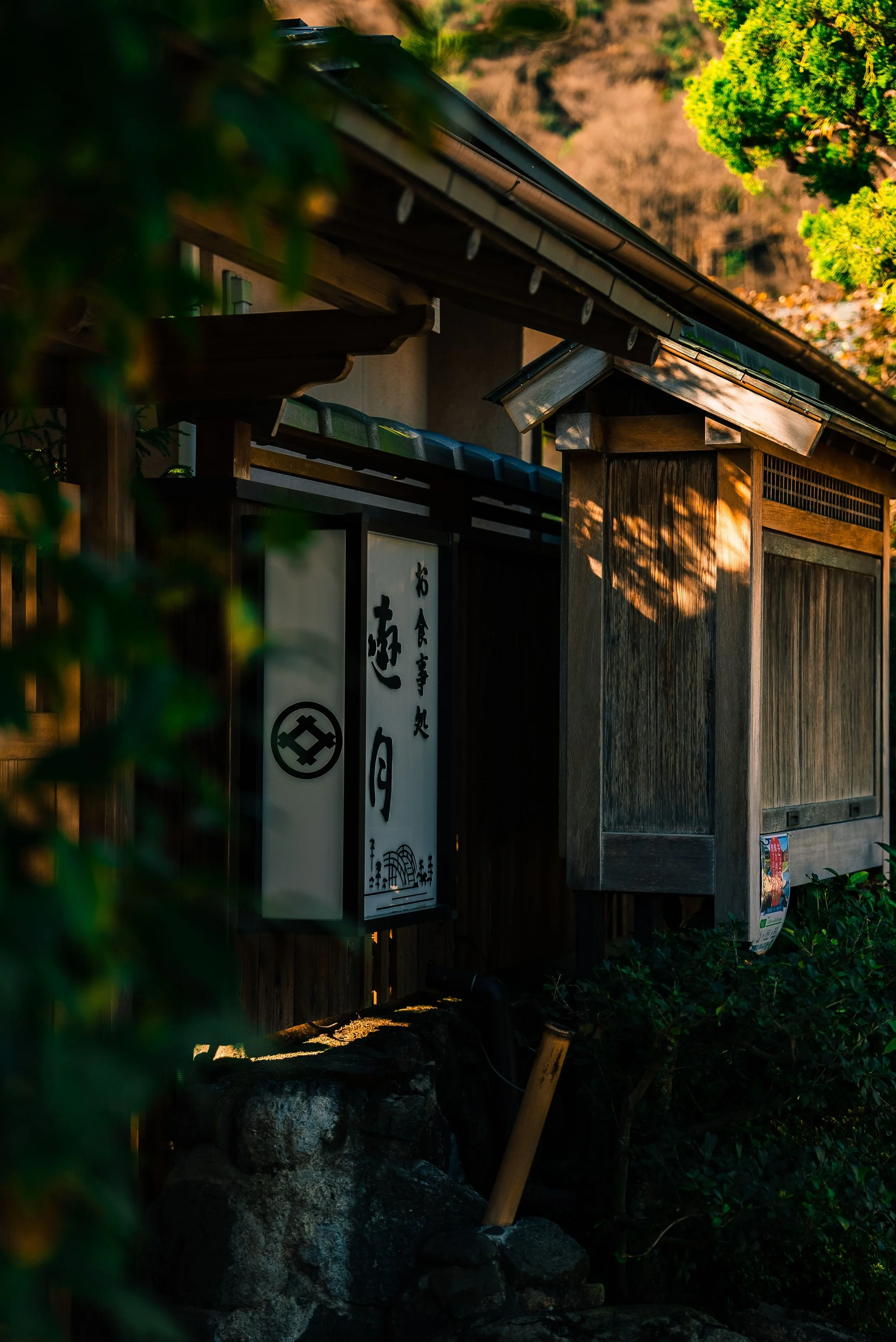A traditional Japanese wooden structure with a tiled roof, partially hidden by green foliage, with a lantern with Japanese characters and a circular emblem on it, set against a background of trees and rocks, illuminated by warm sunlight.