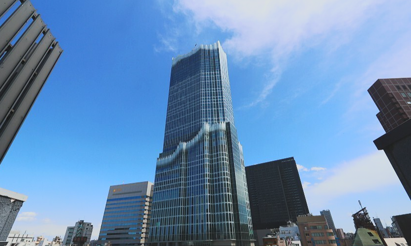 Tall modern glass skyscraper with a unique stepped design in a city skyline, surrounded by other tall buildings under a blue sky with some clouds.