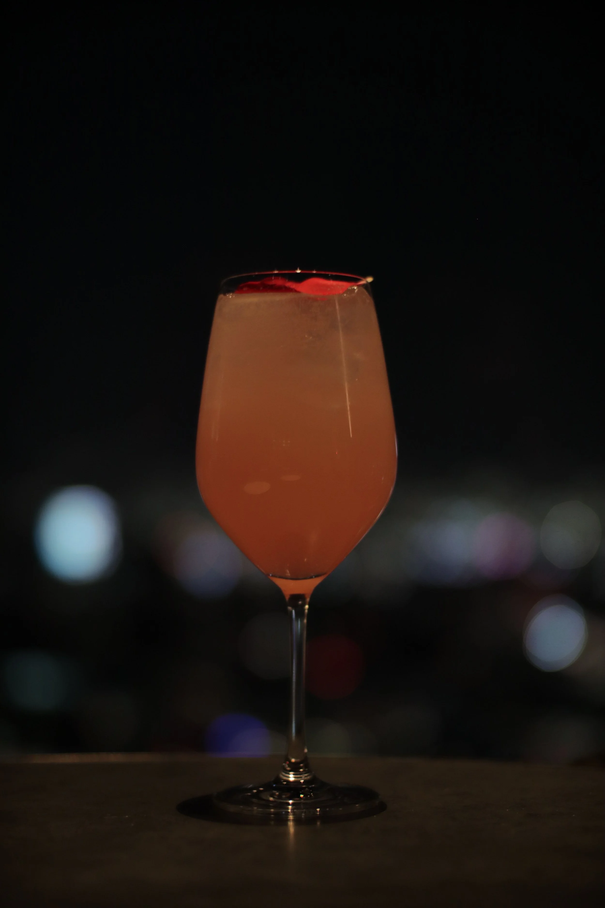 A pink cocktail in a tall, elegant glass with a thin stem, topped with red fruit slices, set against a dark background with blurred city lights.