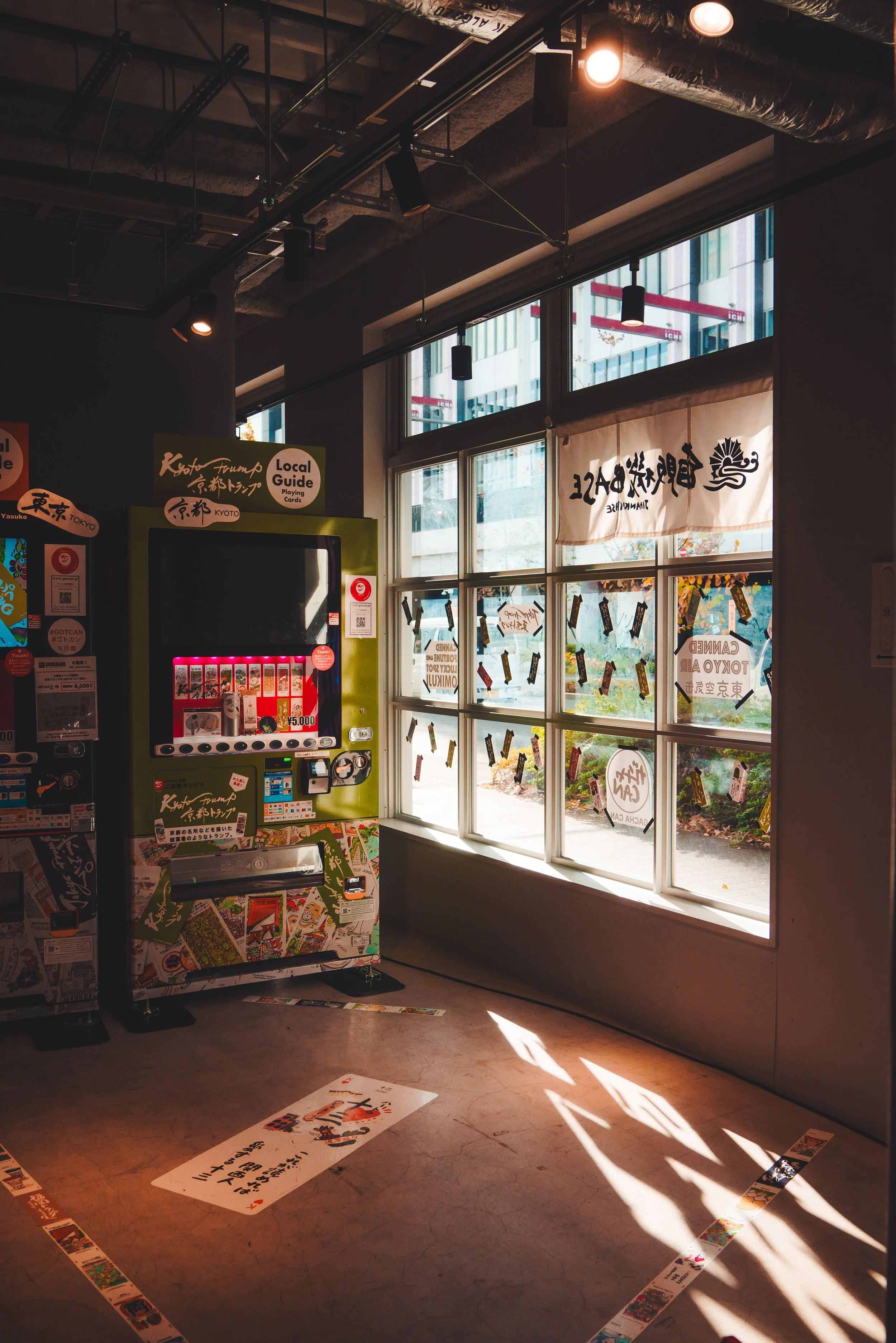 Indoor scene near large windows with sunlight casting shadows on the floor, featuring a Japanese calligraphy banner, a vending machine with colorful buttons, and decorations including playing cards taped to the window and floor.