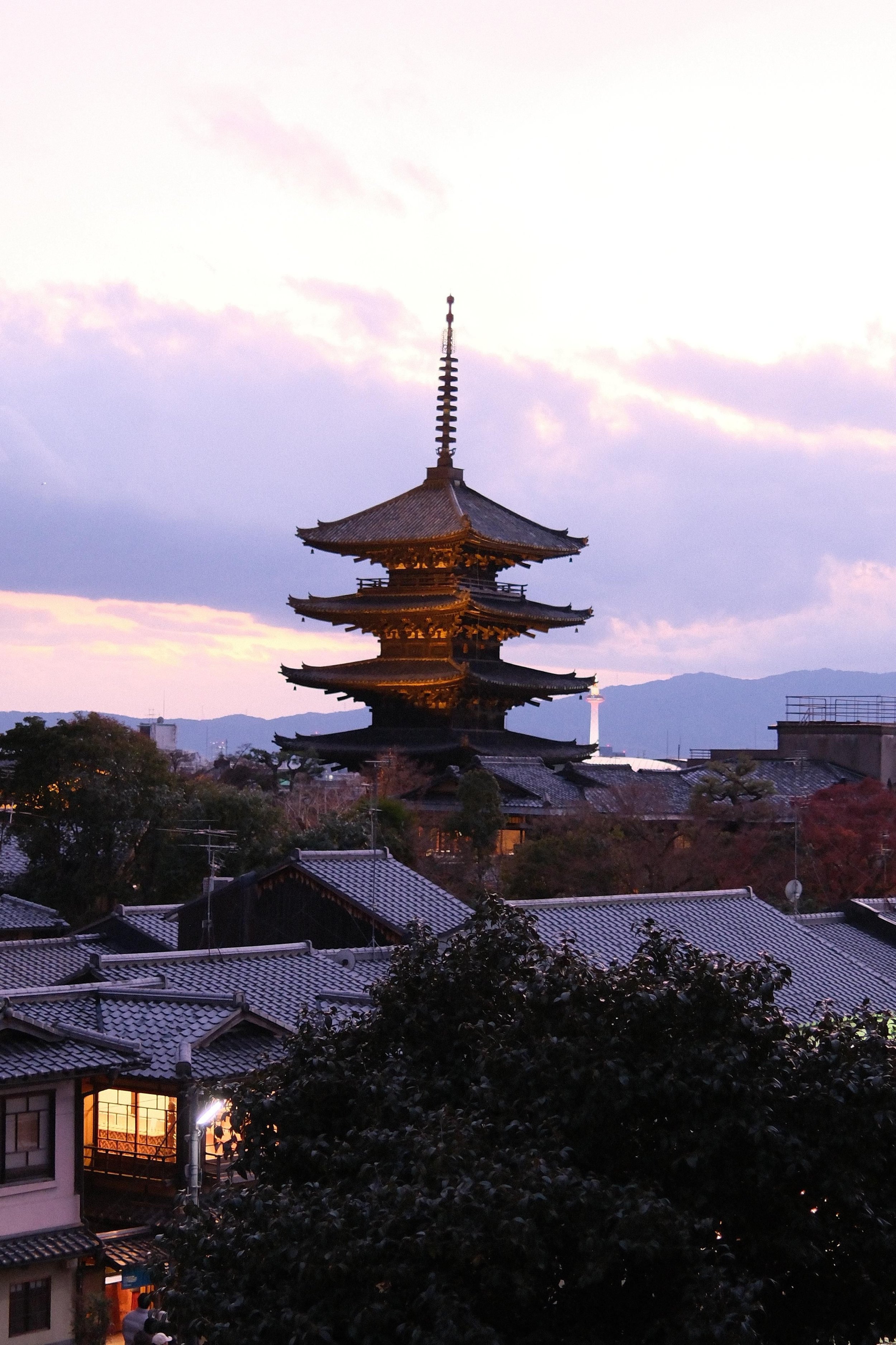 A traditional Japanese five-story pagoda with a mountain range in the background during sunset, with houses and trees in the foreground.