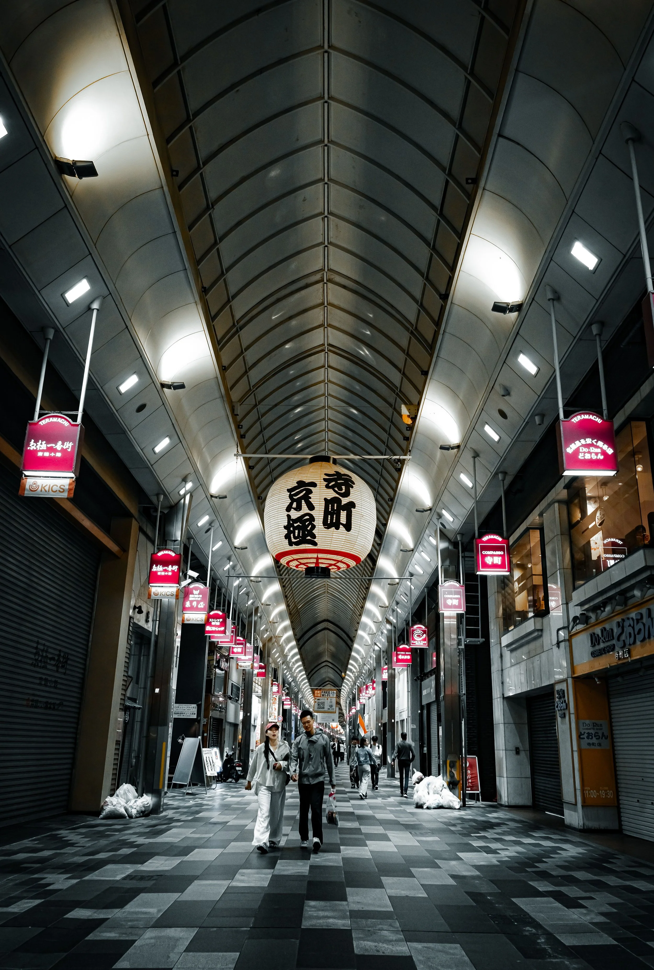 People walking through a covered shopping arcade with a high arched ceiling, illuminated by overhead lights and lanterns with Japanese writing.