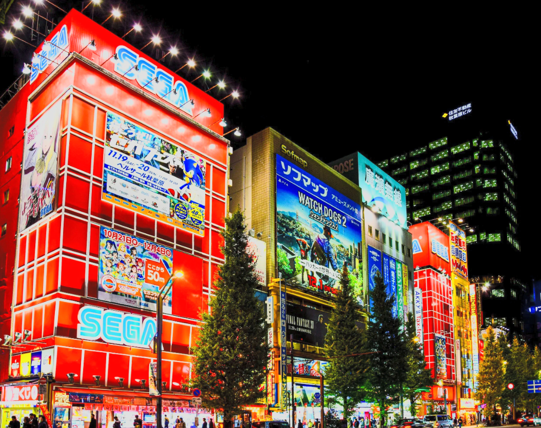 Nighttime city street scene in Tokyo, Japan with brightly lit buildings featuring colorful advertisements and billboards, including prominent SEGA signs and visible storefronts.