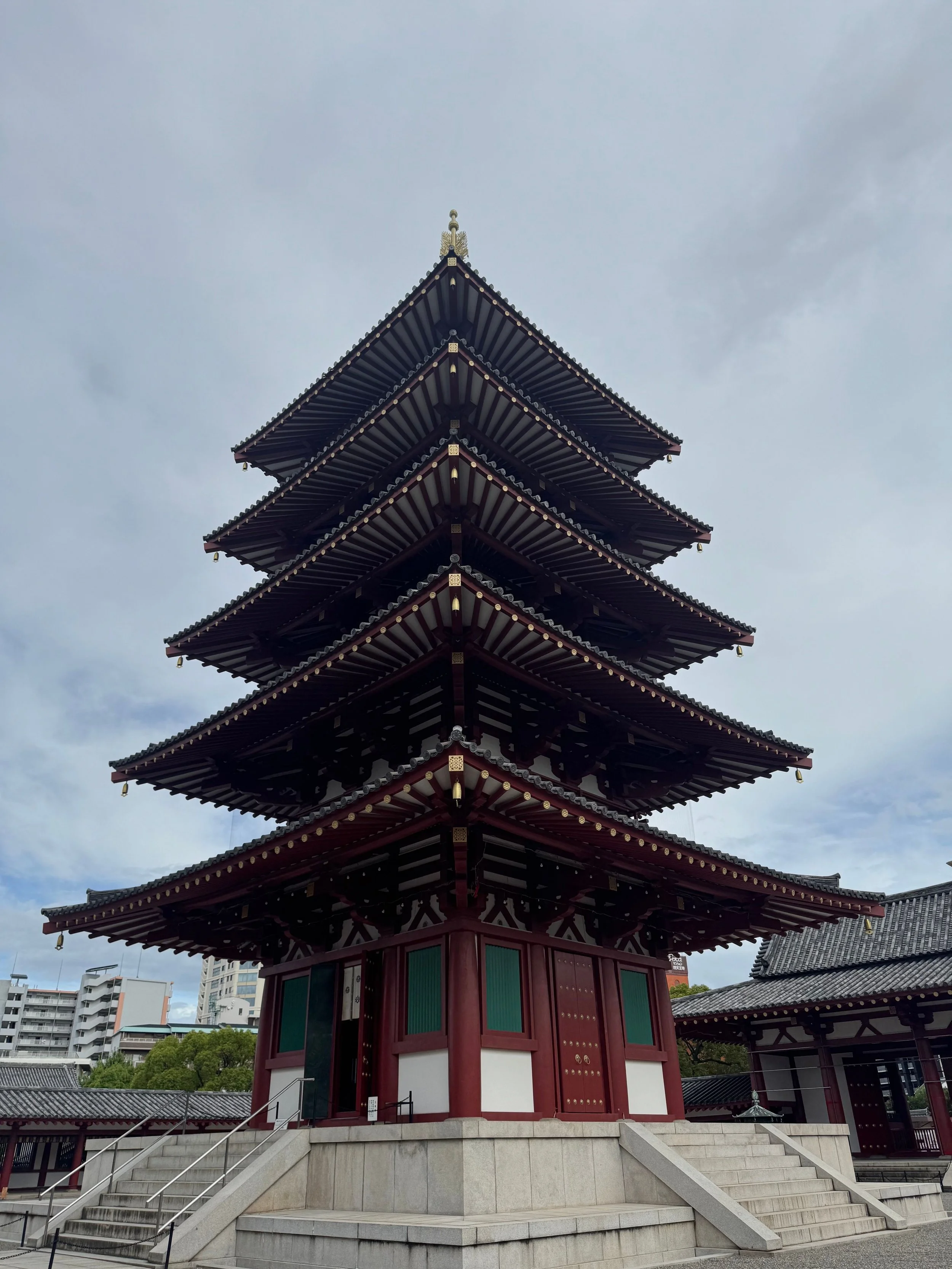 A multi-tiered traditional Japanese pagoda with red and green accents, set on a stone platform with stairs leading up to it. The sky is cloudy, and modern buildings are visible in the background.