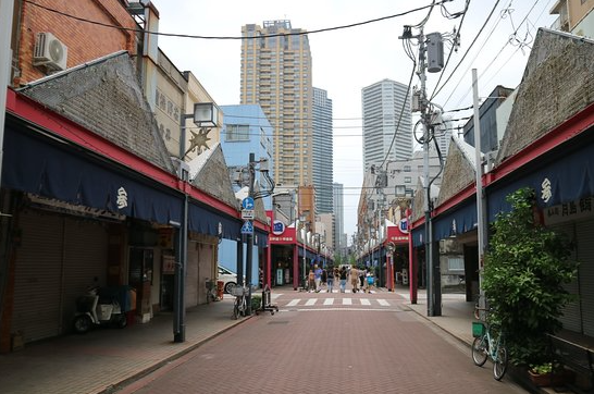 Urban street scene with shops on either side, pedestrians crossing, and tall buildings in the background.