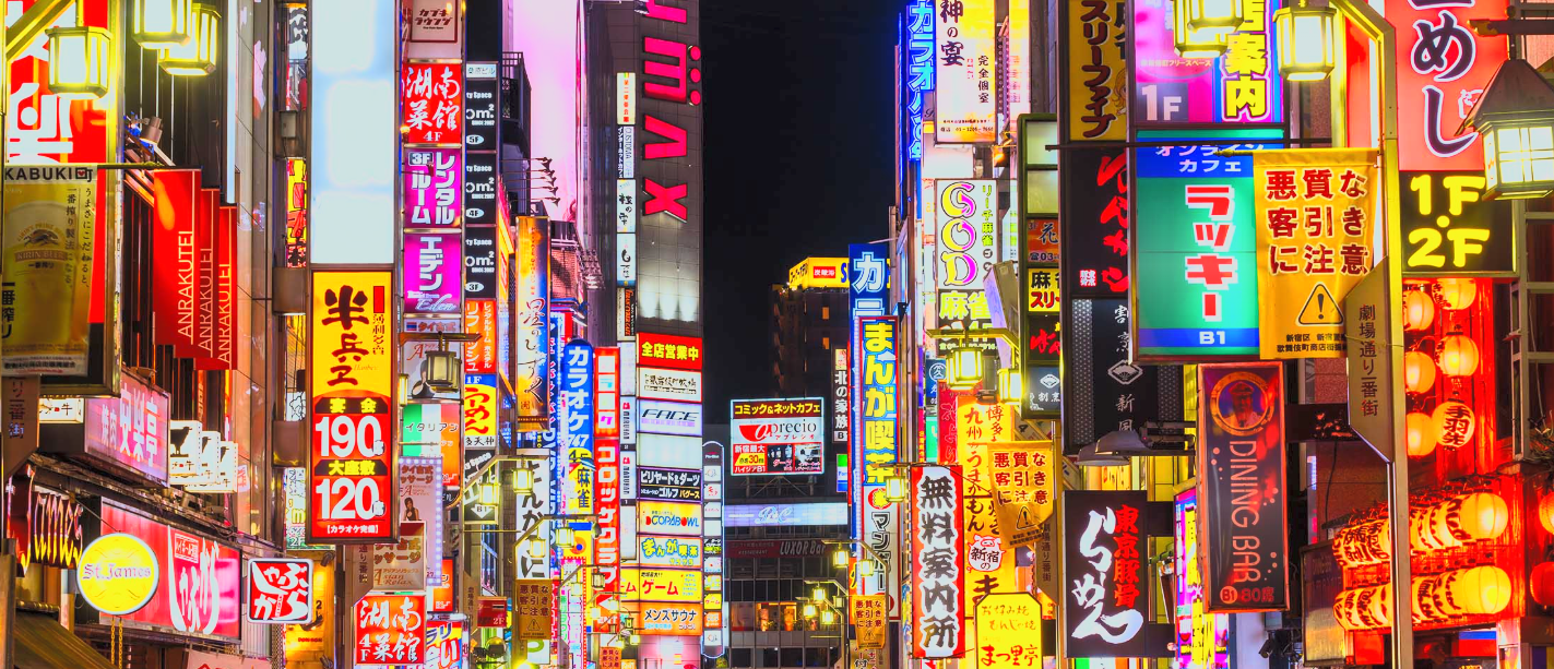 Brightly lit city street at night with numerous colorful neon signs and billboards in Japanese and English, advertising various businesses and products.
