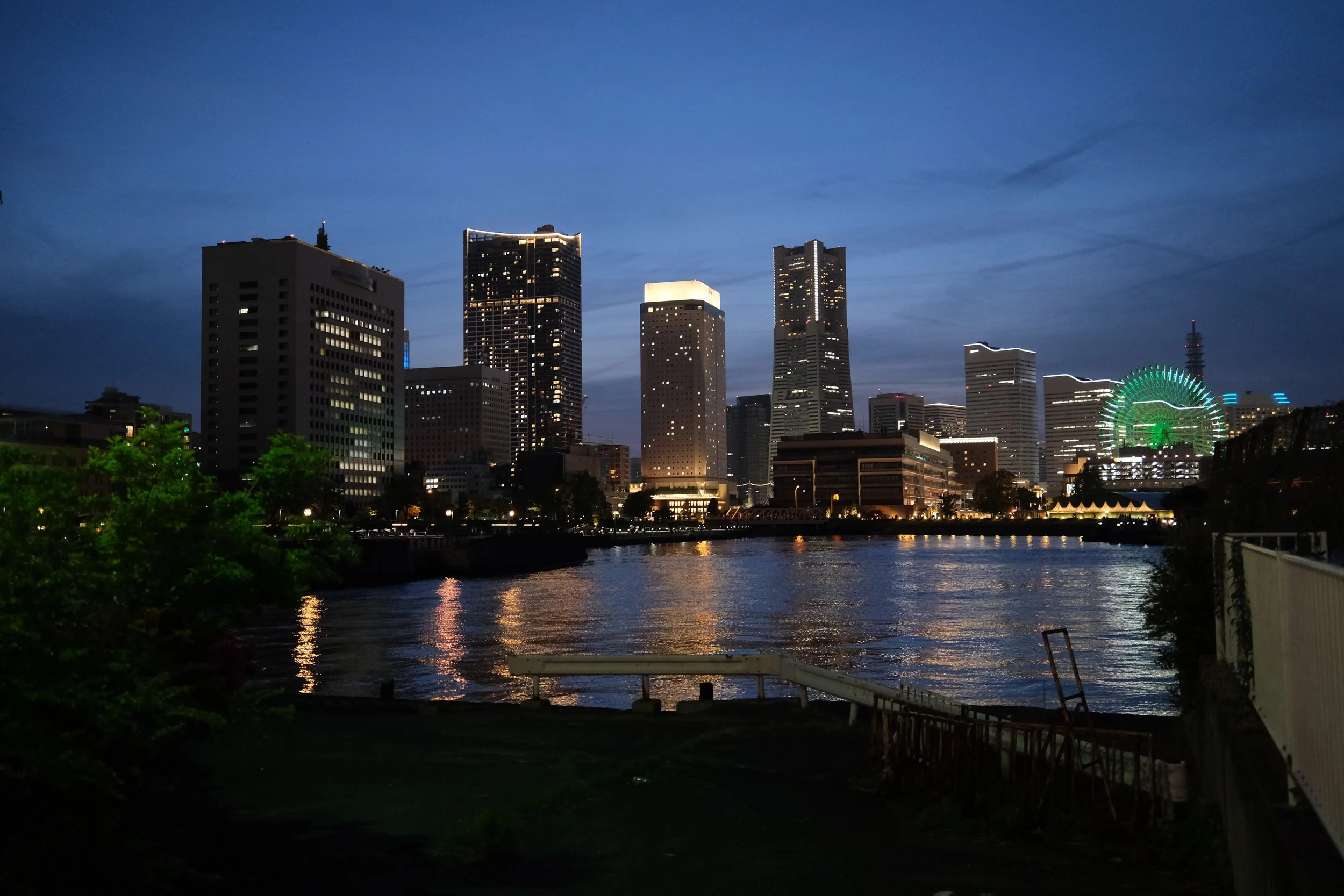 Night skyline of a city with tall buildings illuminated and a Ferris wheel seen in the distance, reflection on a river in the foreground.