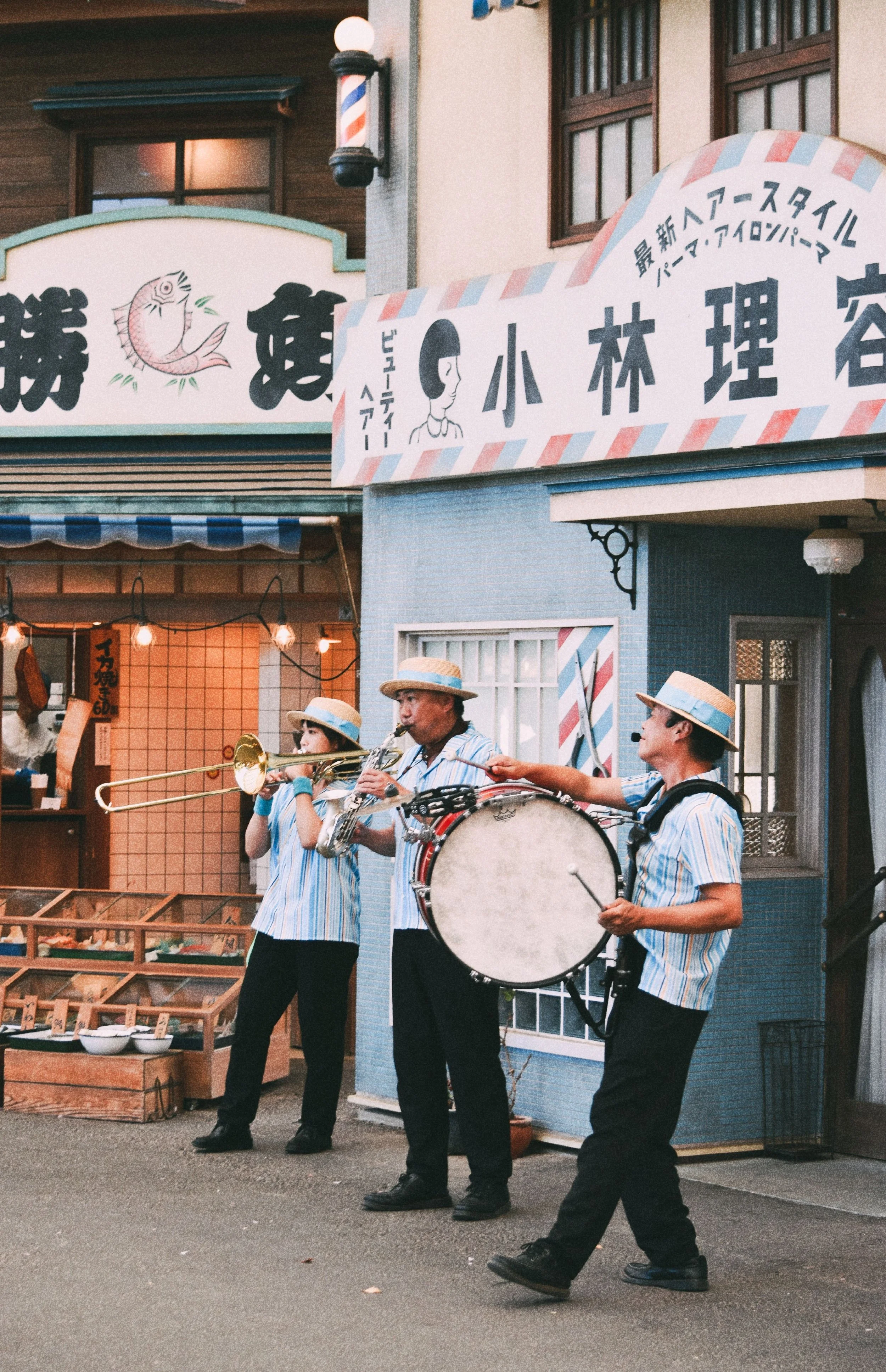 Street scene with three musicians playing instruments, including a bass drum and saxophone, under signage with Japanese characters and a cartoon face, in front of a shop with various items displayed.