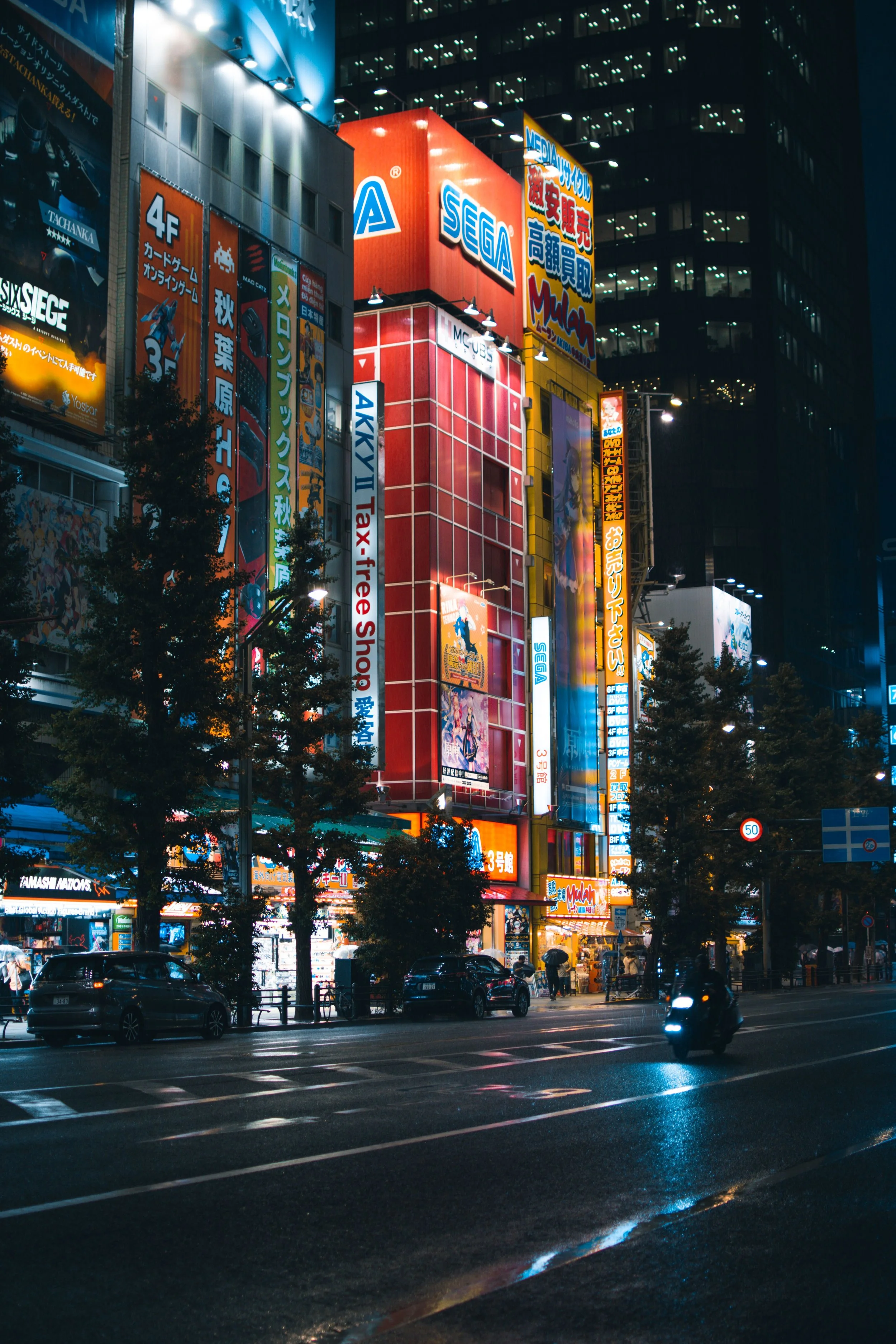 Night view of a busy city street with illuminated buildings covered in colorful advertisements and signs. Cars are driving along the wet road, and pedestrians are walking on the sidewalk under umbrellas.