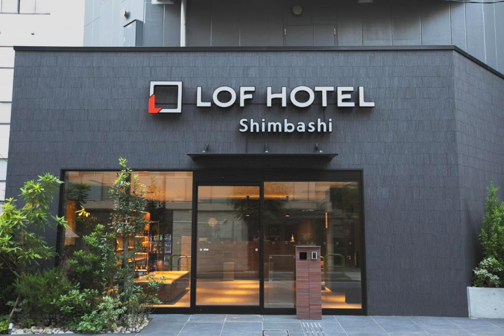 Exterior view of the LOF Hotel Shimbashi, showing the hotel entrance with large glass doors, greenery on either side, and a sign displaying the hotel name and location.