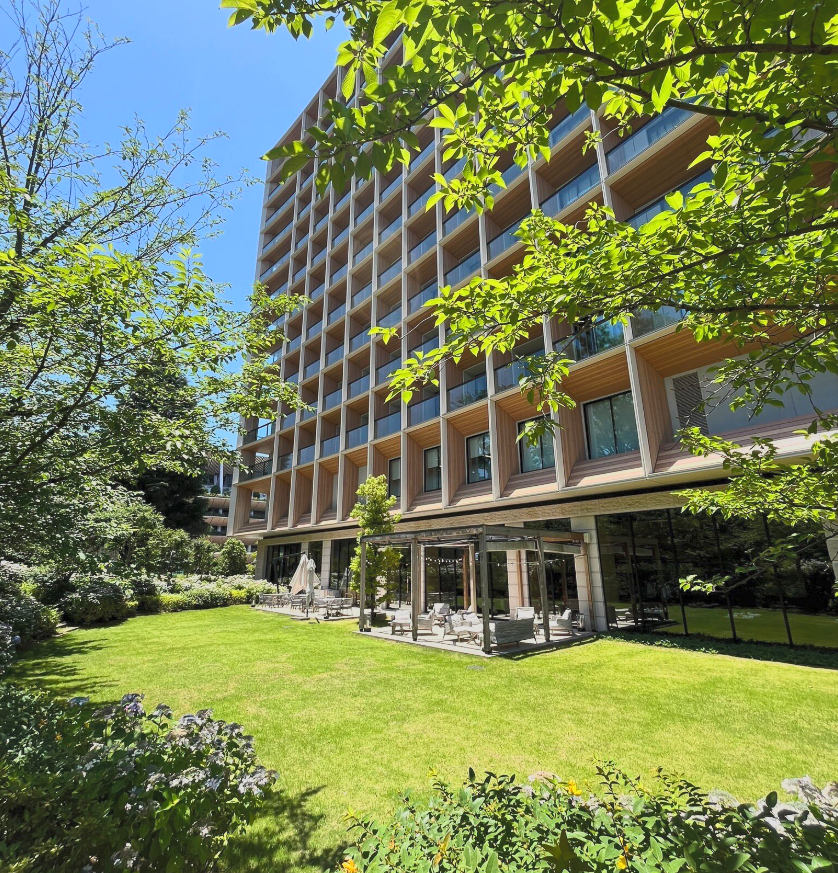 A modern multi-story building with a unique architectural facade surrounded by greenery, including trees and a well-maintained lawn, with outdoor seating and a patio area in the foreground.