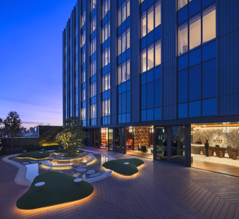 Modern hotel building with glowing windows and a rooftop garden patio with landscaped greenery at dusk.