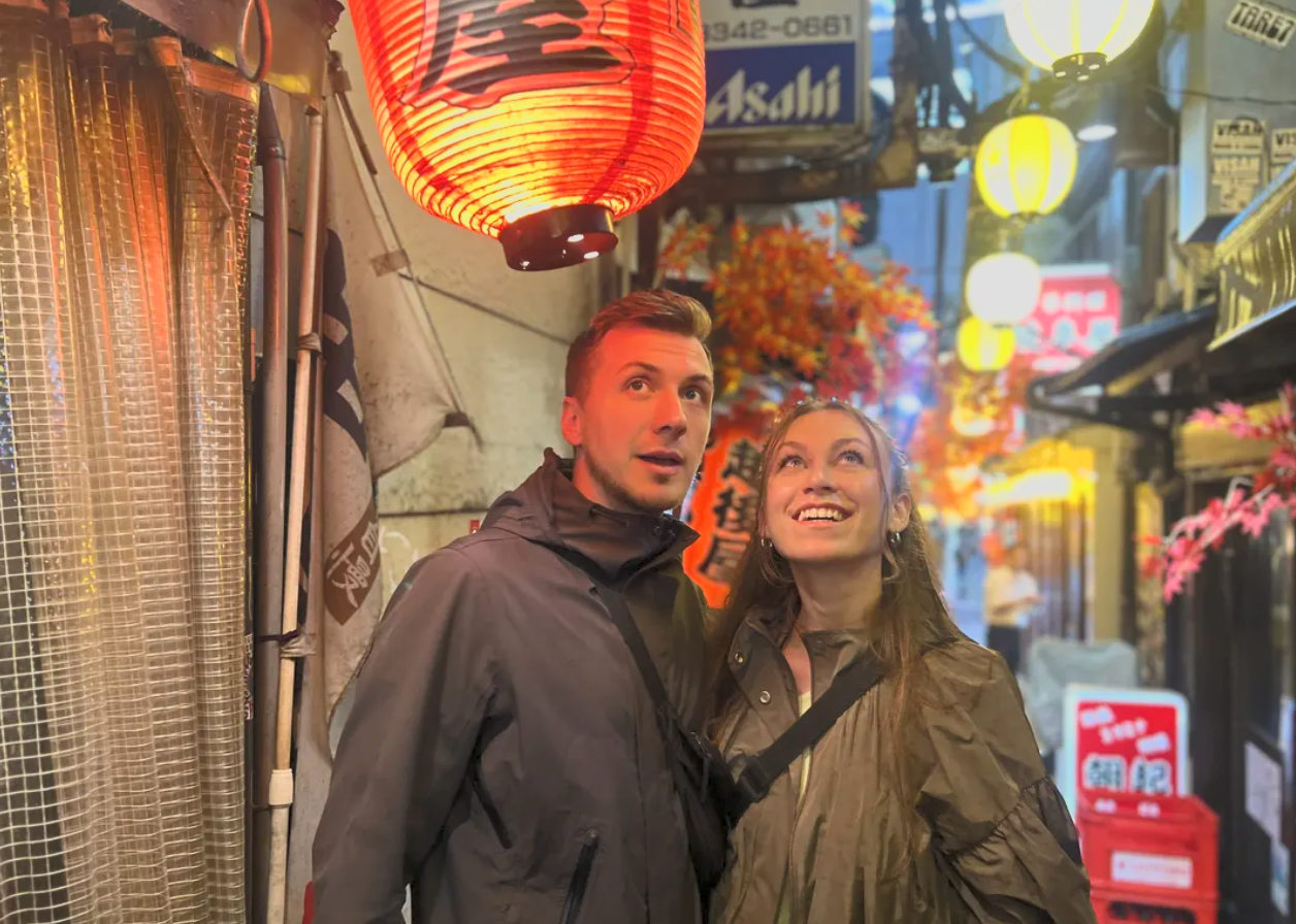A young man and woman standing close together in a vibrant, illuminated street filled with lanterns, signs, and colorful decorations, likely in an Asian city at night.