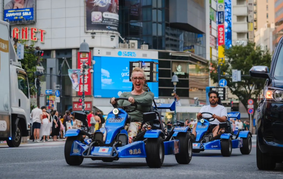 Two people riding toy cars on a busy city street with onlookers and tall buildings background.