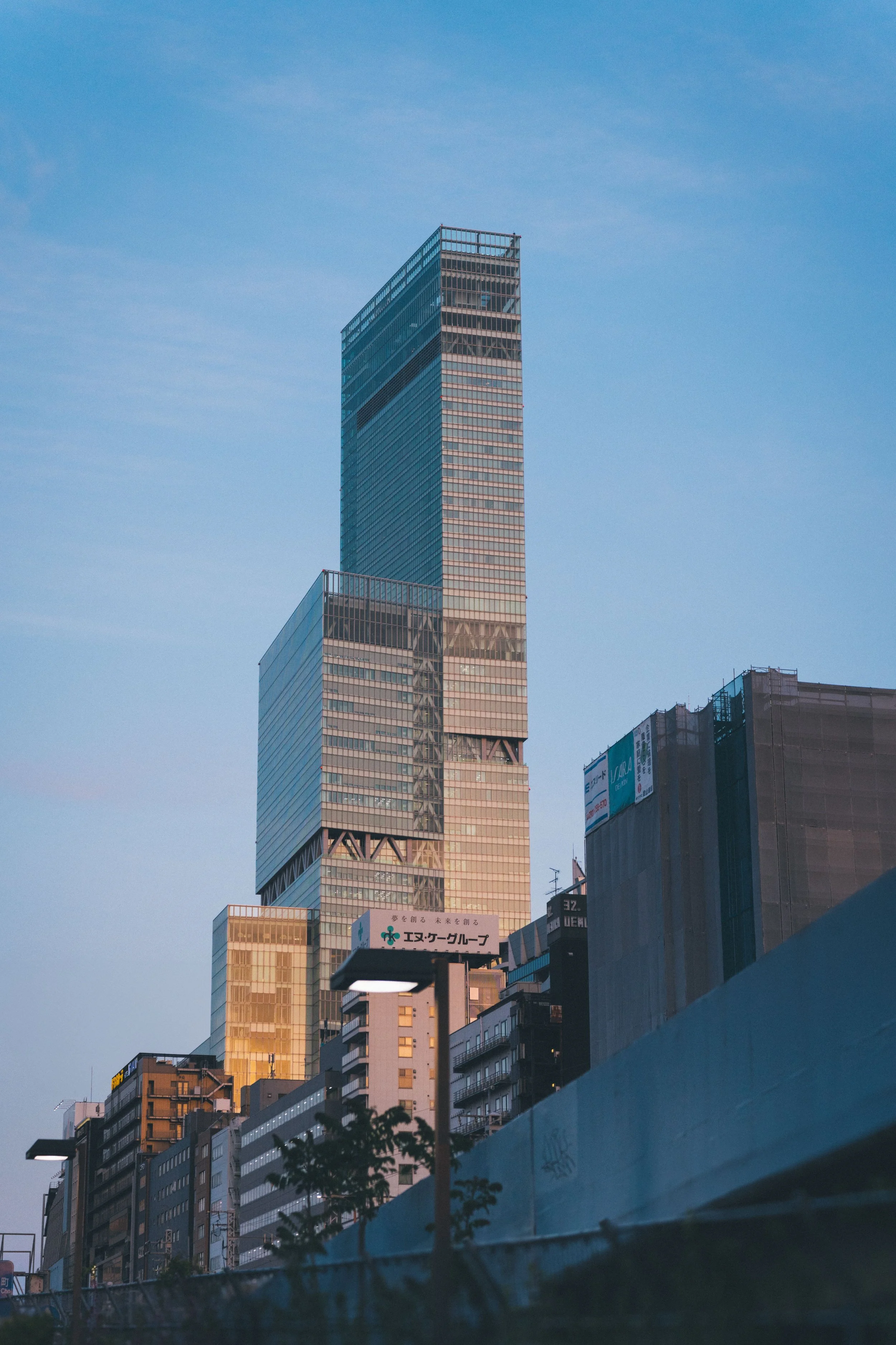 A cityscape featuring tall modern skyscrapers under a clear blue sky, with buildings illuminated by sunlight, and a few trees at the bottom.