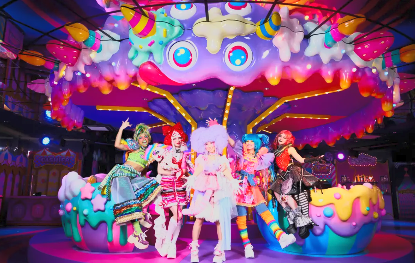 Colorful, whimsical carousel with five performers dressed in vibrant, playful costumes, surrounded by oversized candy-themed decorations.
