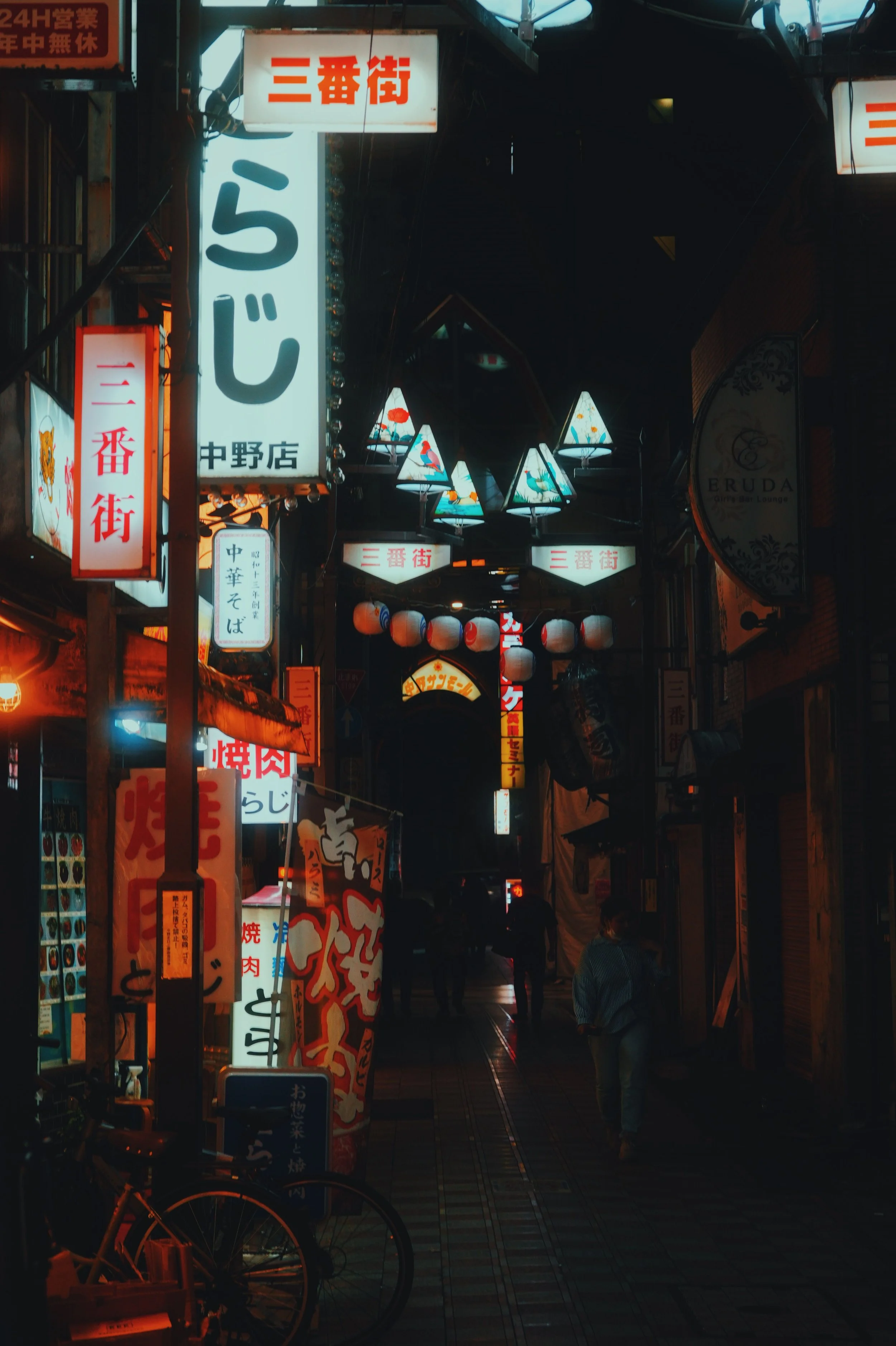 Night street scene in Japan with illuminated signs in Japanese, paper lanterns, and triangular lamps hanging overhead. Several pedestrians are walking along the sidewalk.