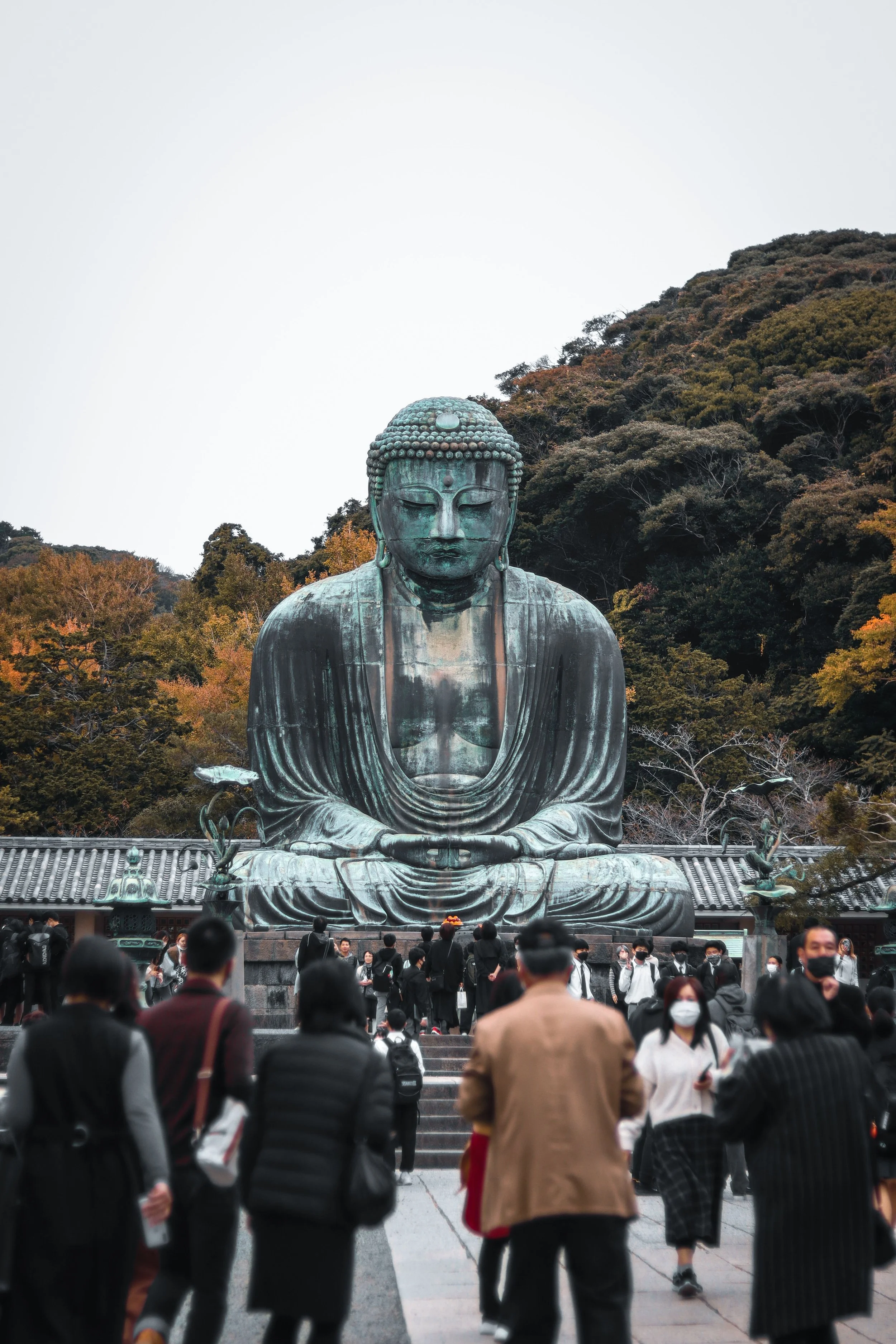 Large bronze statue of Buddha in sitting position with crossed legs, hands resting on lap, surrounded by a crowd of people, with trees and hillside in the background.