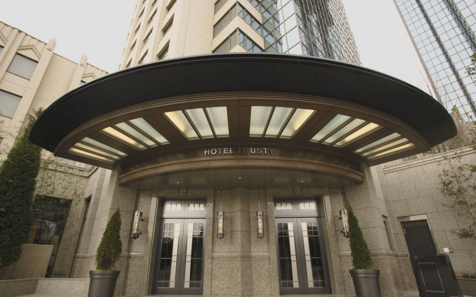 Exterior view of Hotel Trusty with a rounded black canopy, glass double doors, and potted plants outside in an urban setting.