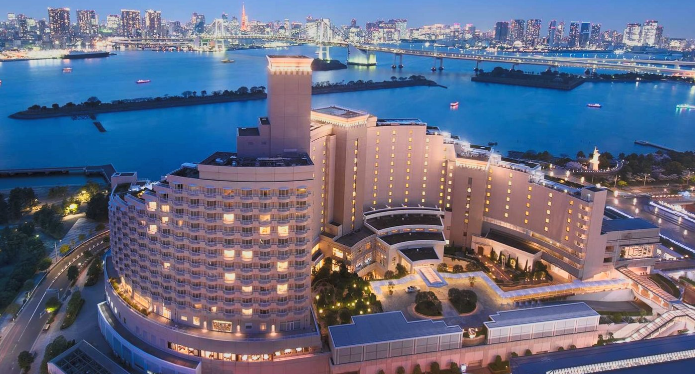 Aerial view of a large, illuminated hotel or resort overlooking a river at dusk, with a city skyline in the background.
