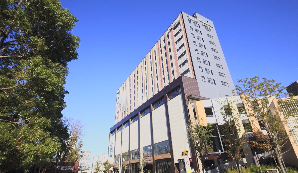 A tall modern hotel building with multiple floors and a mix of windows and exterior panels, with trees in the foreground and a clear blue sky in the background.