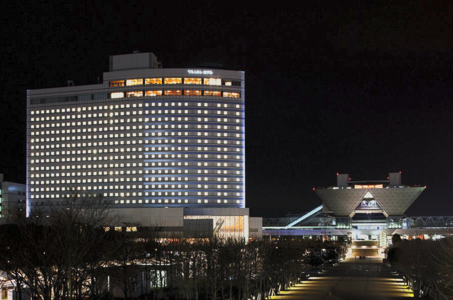 Night view of a tall hotel building with illuminated windows and a distinctive modern conference center or stadium in the background, with trees and a pathway in the foreground.