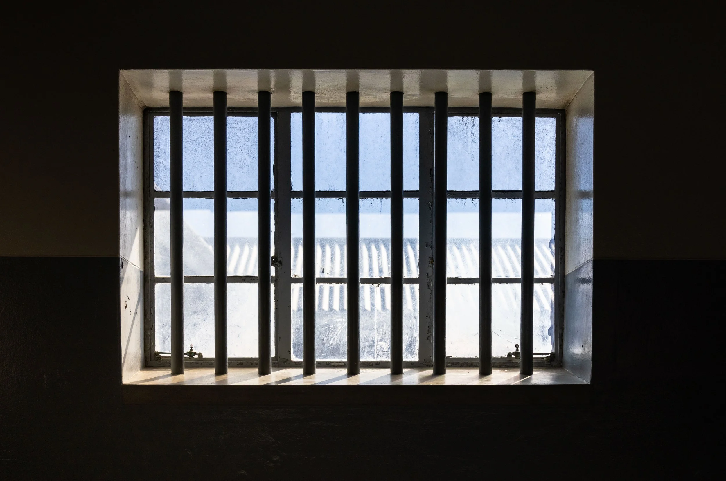A barred jail cell window with frosted glass and metal bars, viewed from inside a dark room.