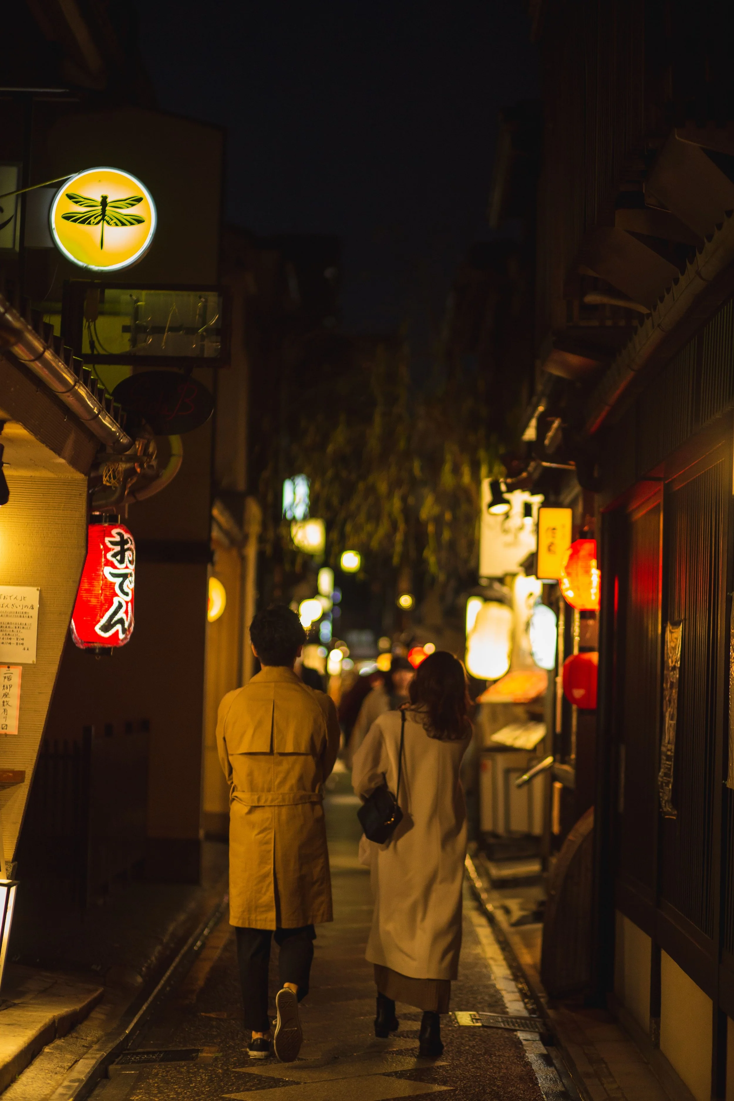 Night scene of a busy street in Japan with people walking, illuminated signs, and a dragonfly logo sign on a building