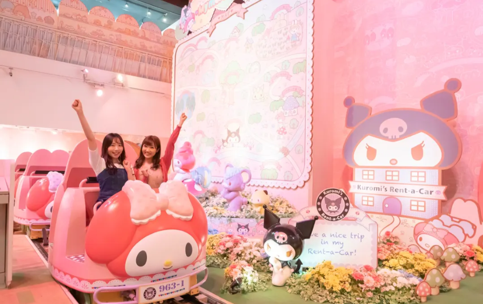 Two women cheerfully sitting in pink ride-on cars themed with Sanrio characters, in a colorful, decorated indoor setting with pink flowers and plush toys, featuring large character signs and a screen with a message.