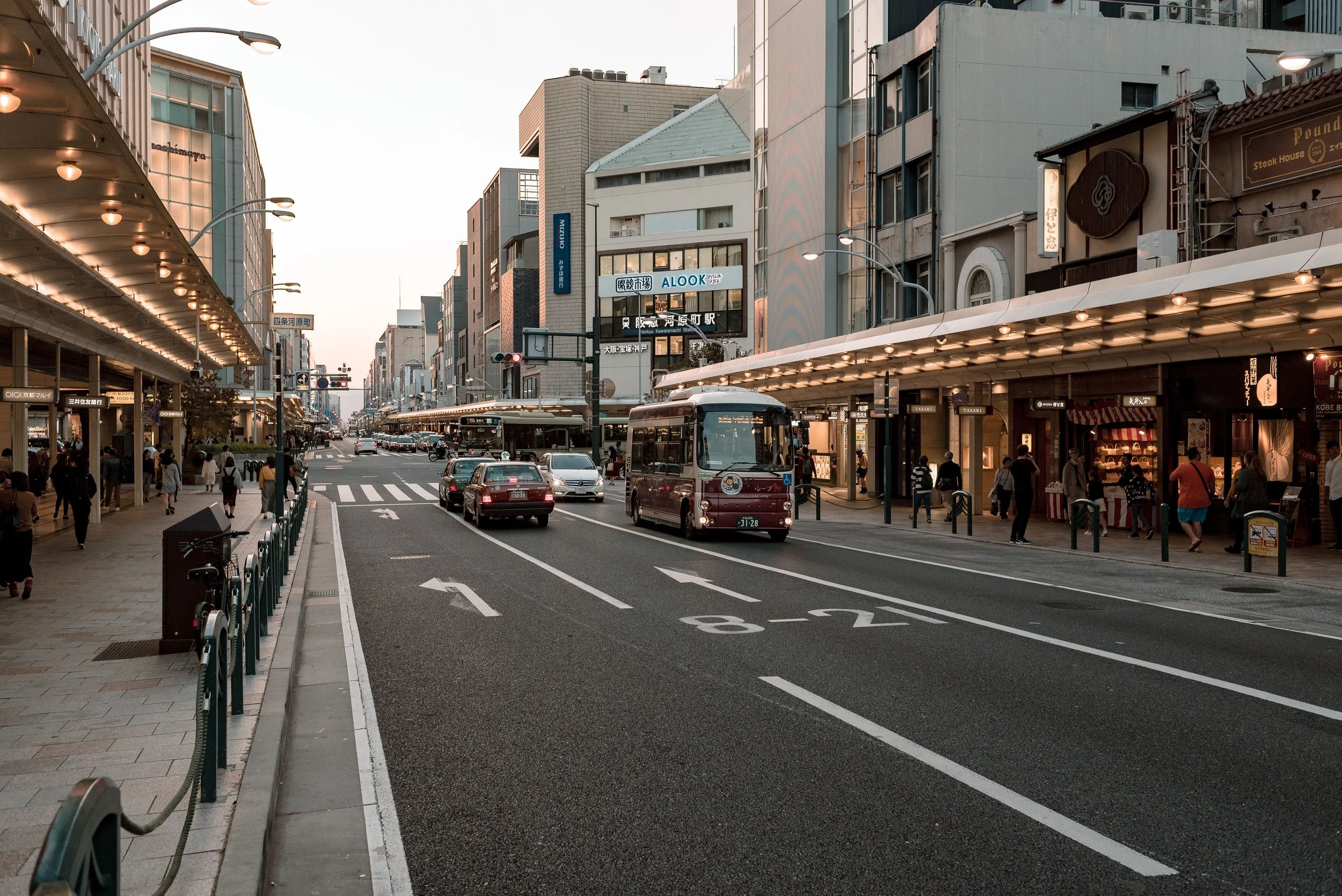 A busy city street in Japan with pedestrians walking on the sidewalks, vehicles on the road, and storefronts with signs and lights, during early evening or dusk.