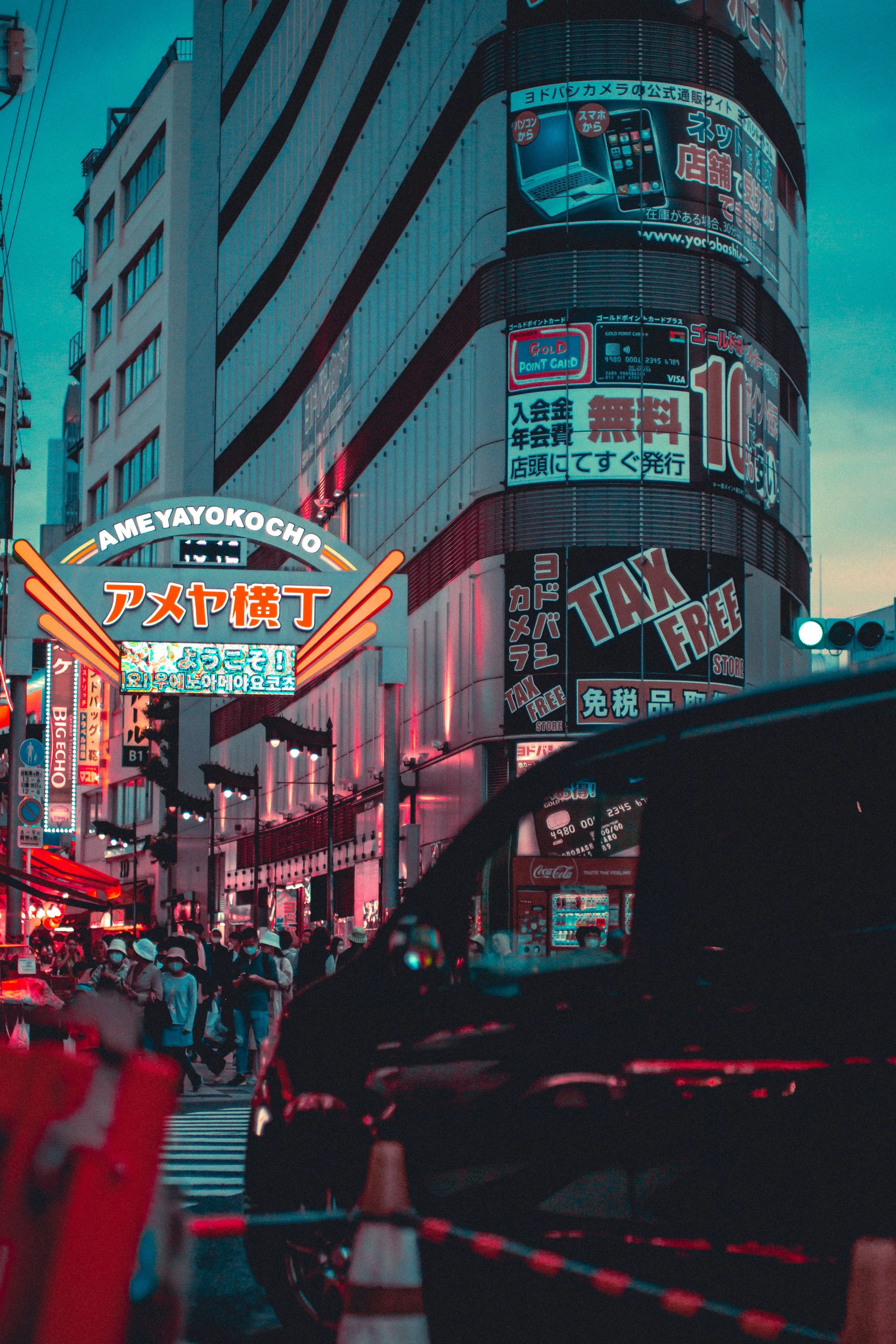 A busy downtown street scene in the evening, illuminated by colorful neon signs in Japanese, with a crowd of pedestrians crossing the street and buildings adorned with advertisements and digital billboards.
