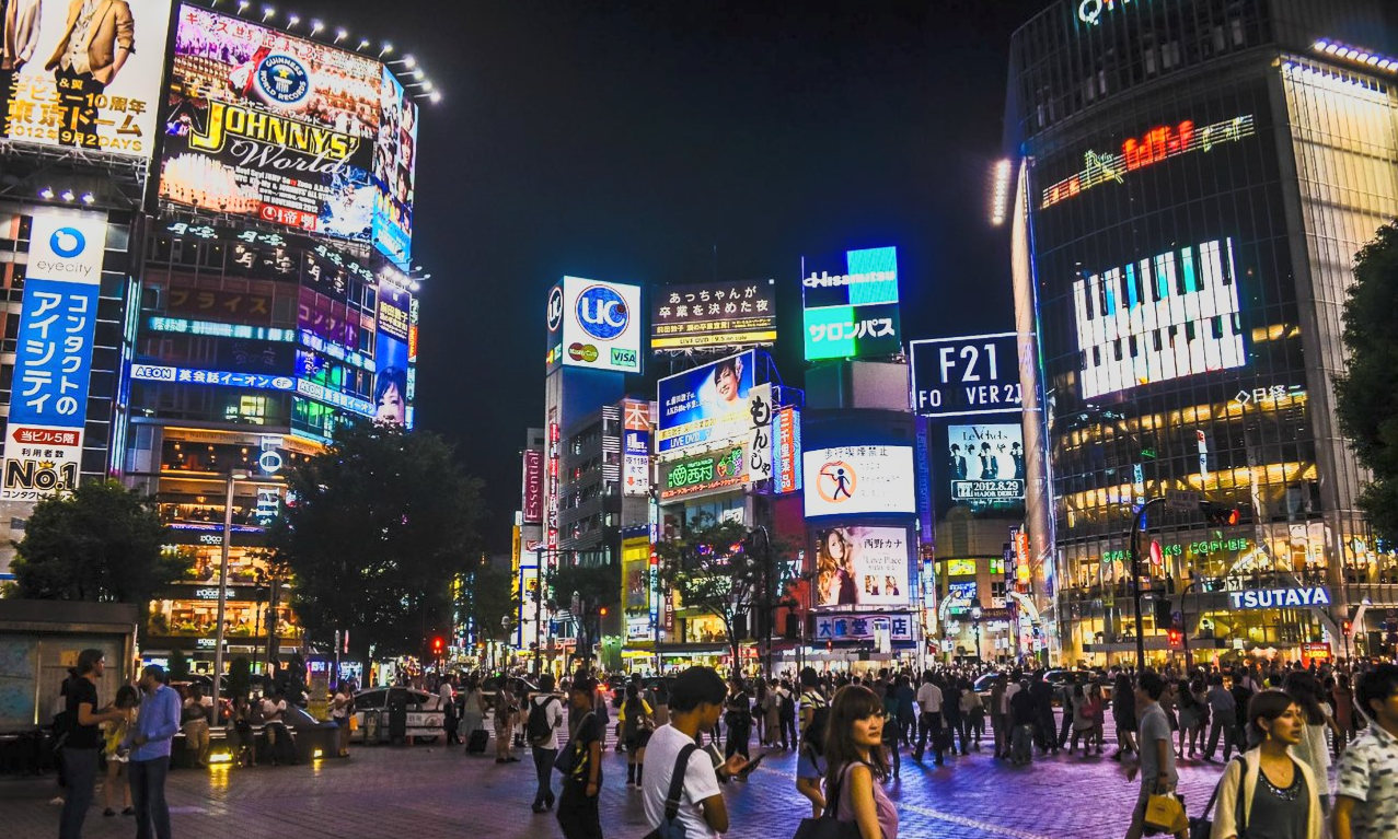 Night scene of a busy city intersection with bright billboards and neon signs, crowds of people walking, and tall glass buildings in the background, likely in Tokyo's Shibuya district.